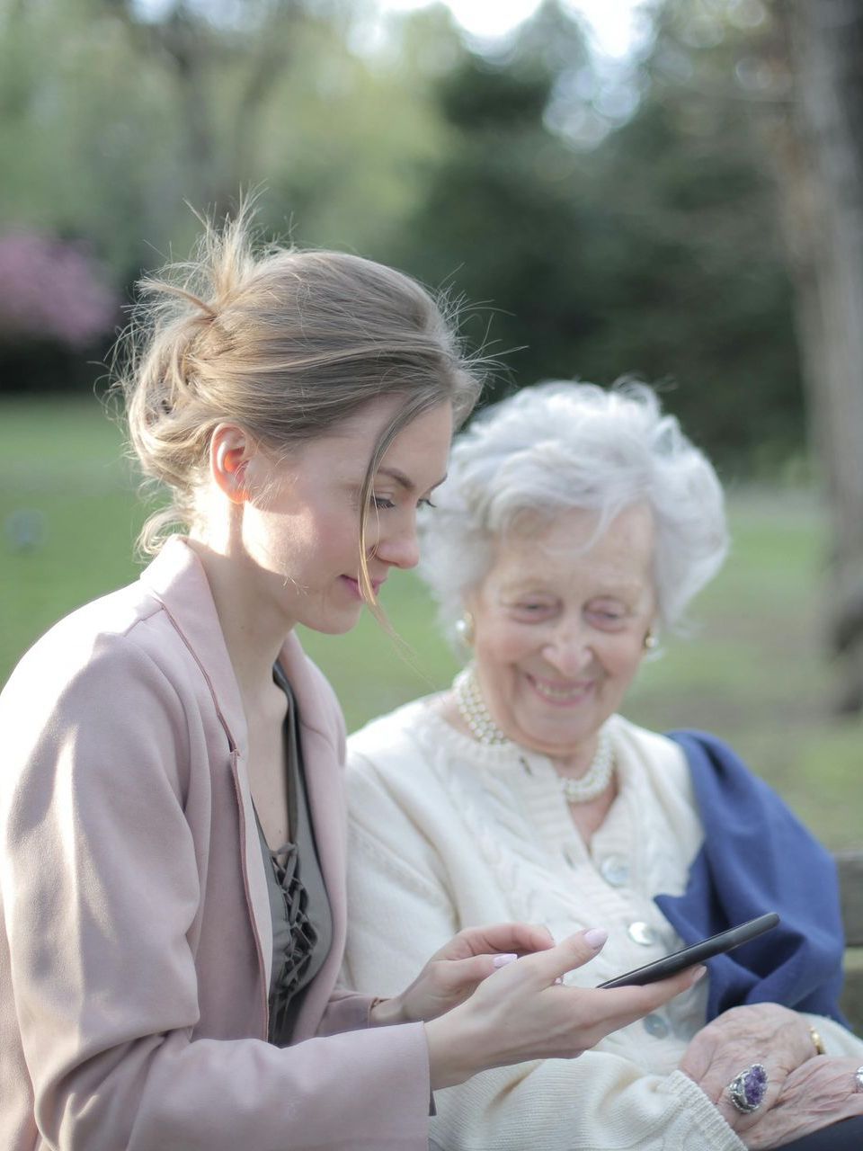 Two women are sitting on a bench in a park looking at a cell phone.