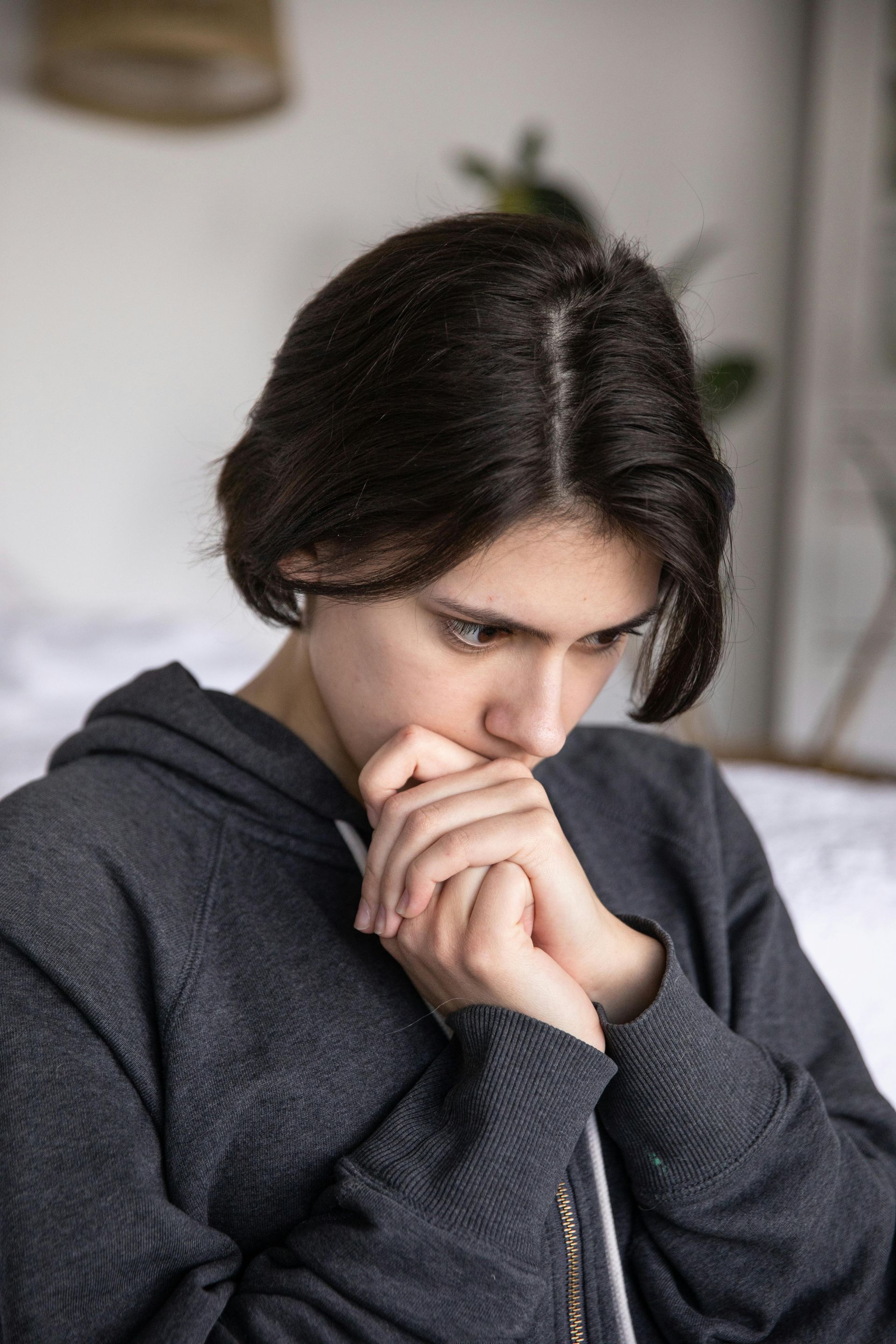 Person in a gray hoodie with a concerned expression, hands clasped near face.