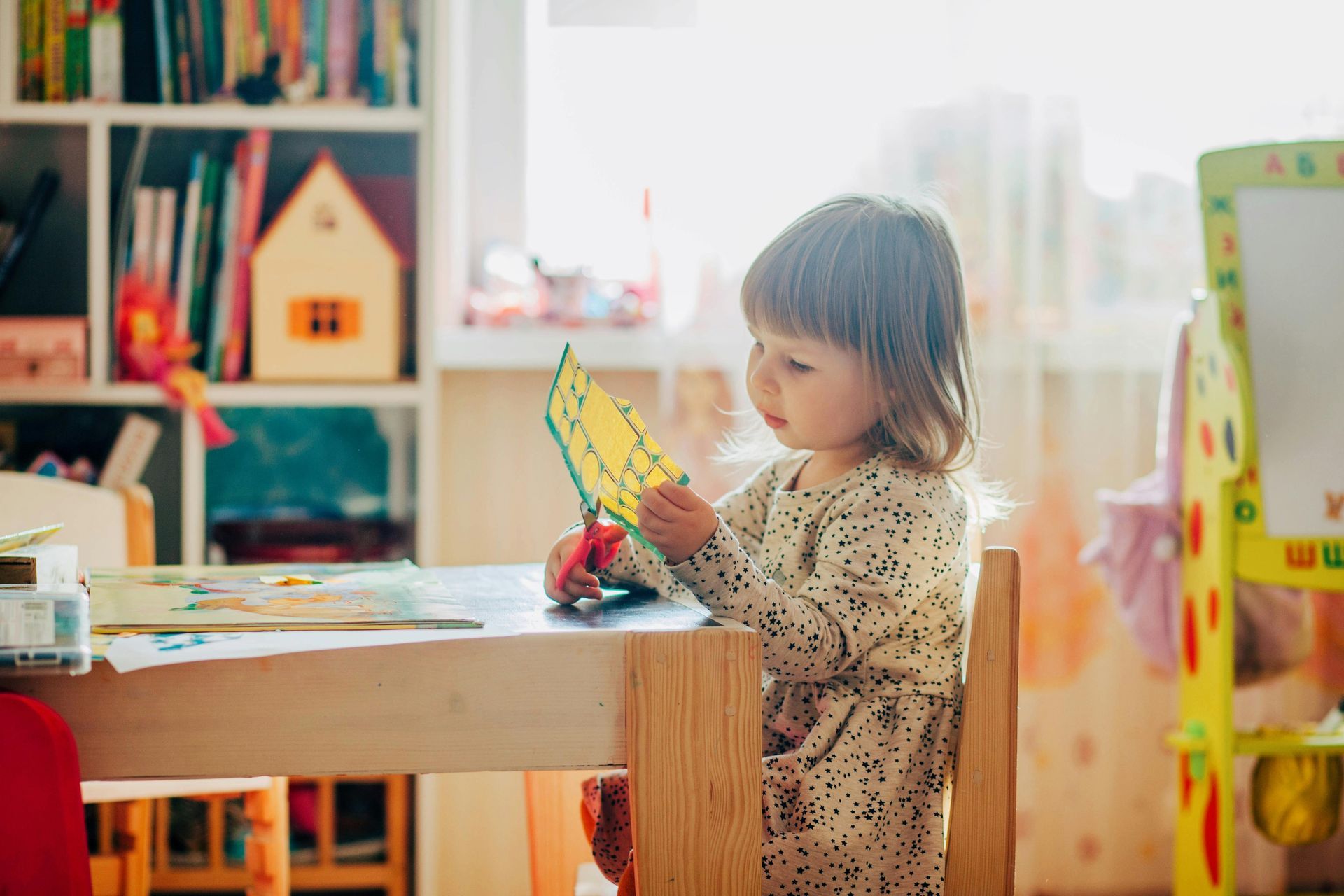 A young child with short hair cuts yellow paper with scissors at a table near a bookcase.