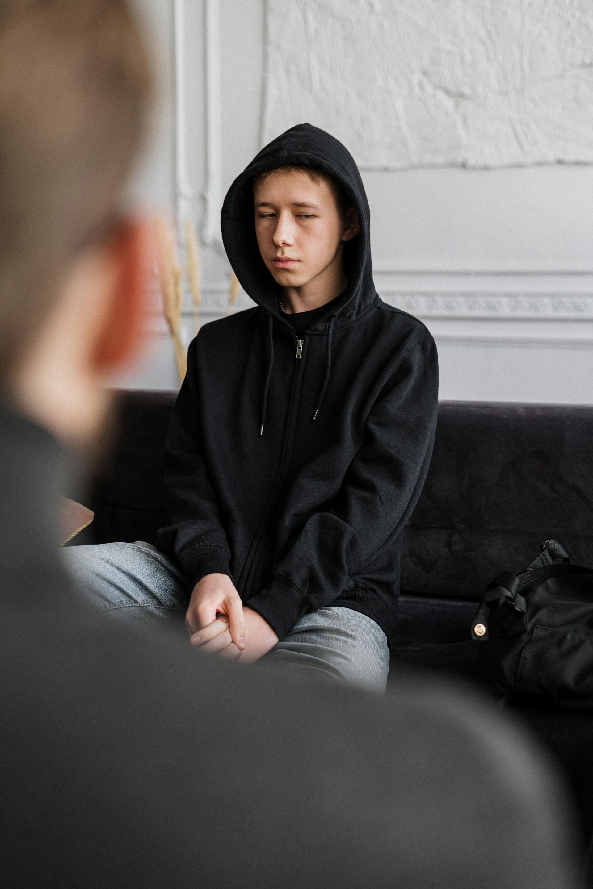 Young person in black hoodie with eyes closed, sitting on a couch, facing another person out of focus.