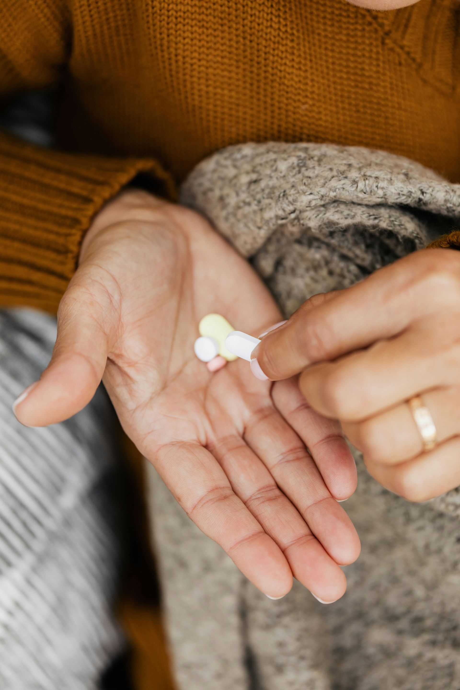 Person holding pills in their open palm, some of them are colored. They're wearing a brown sweater and covered with a gray blanket.