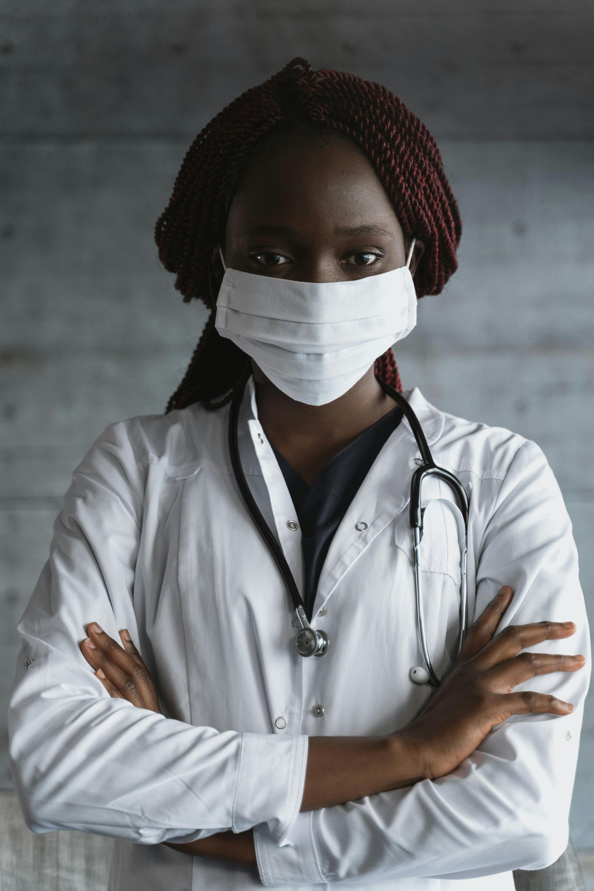A female doctor wearing a mask and a stethoscope is standing with her arms crossed.