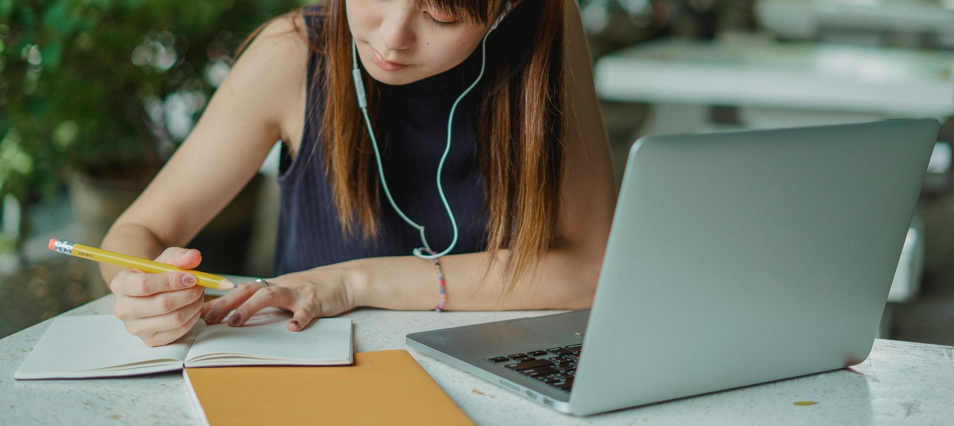 Girl writing in a notebook, using a laptop and listening with earphones.