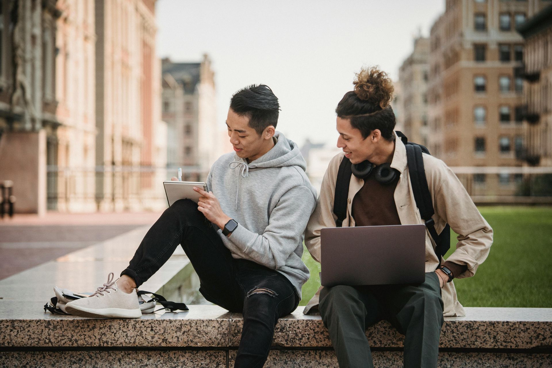 Two students sit outdoors, one with a tablet, one with a laptop, smiling.