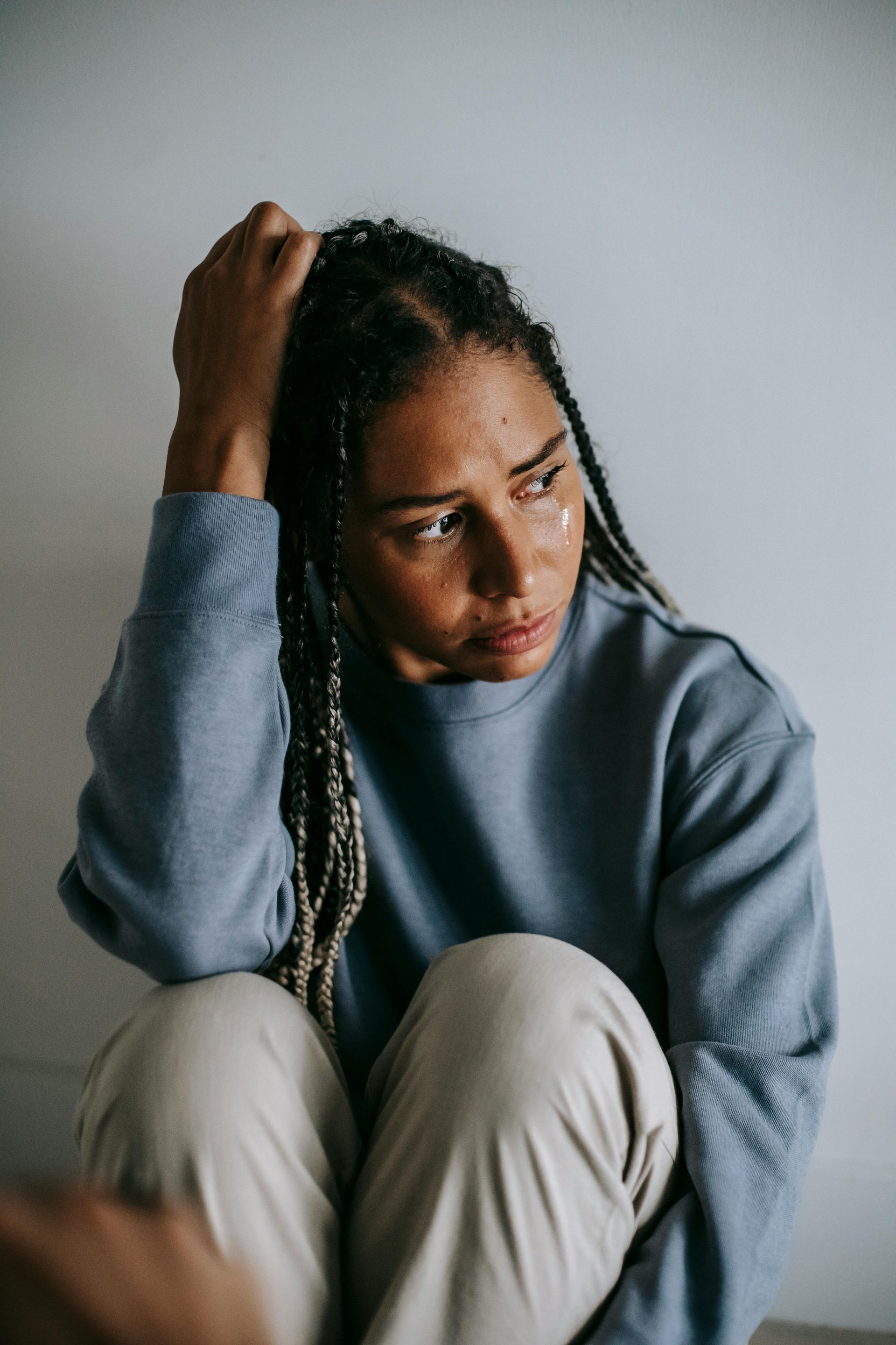 A woman with braided hair sits with a worried expression, hand on her head, looking away. She wears a blue sweater and light-colored pants, against a white wall.