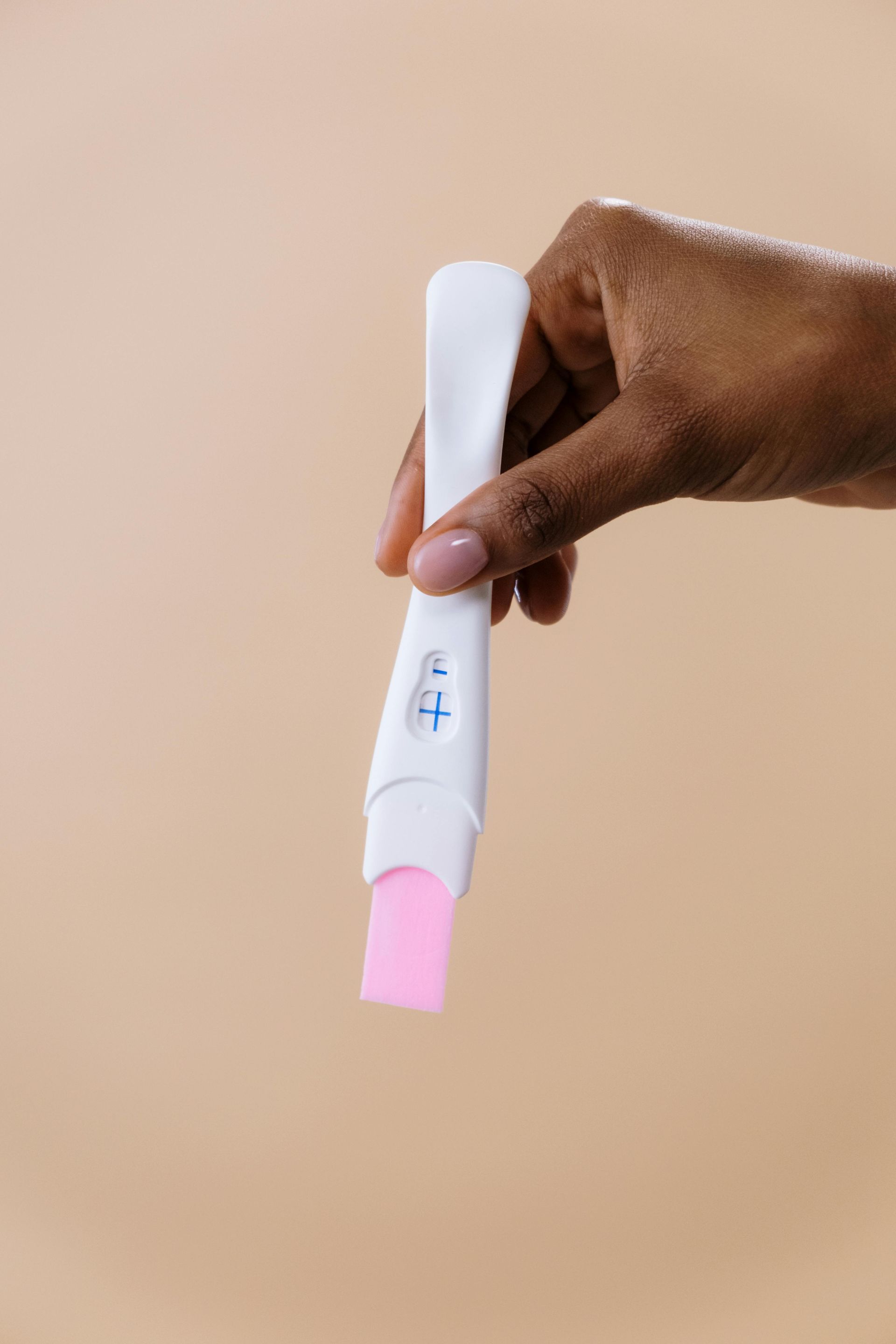 A hand holds a white pregnancy test showing a positive blue plus sign against a plain beige background.