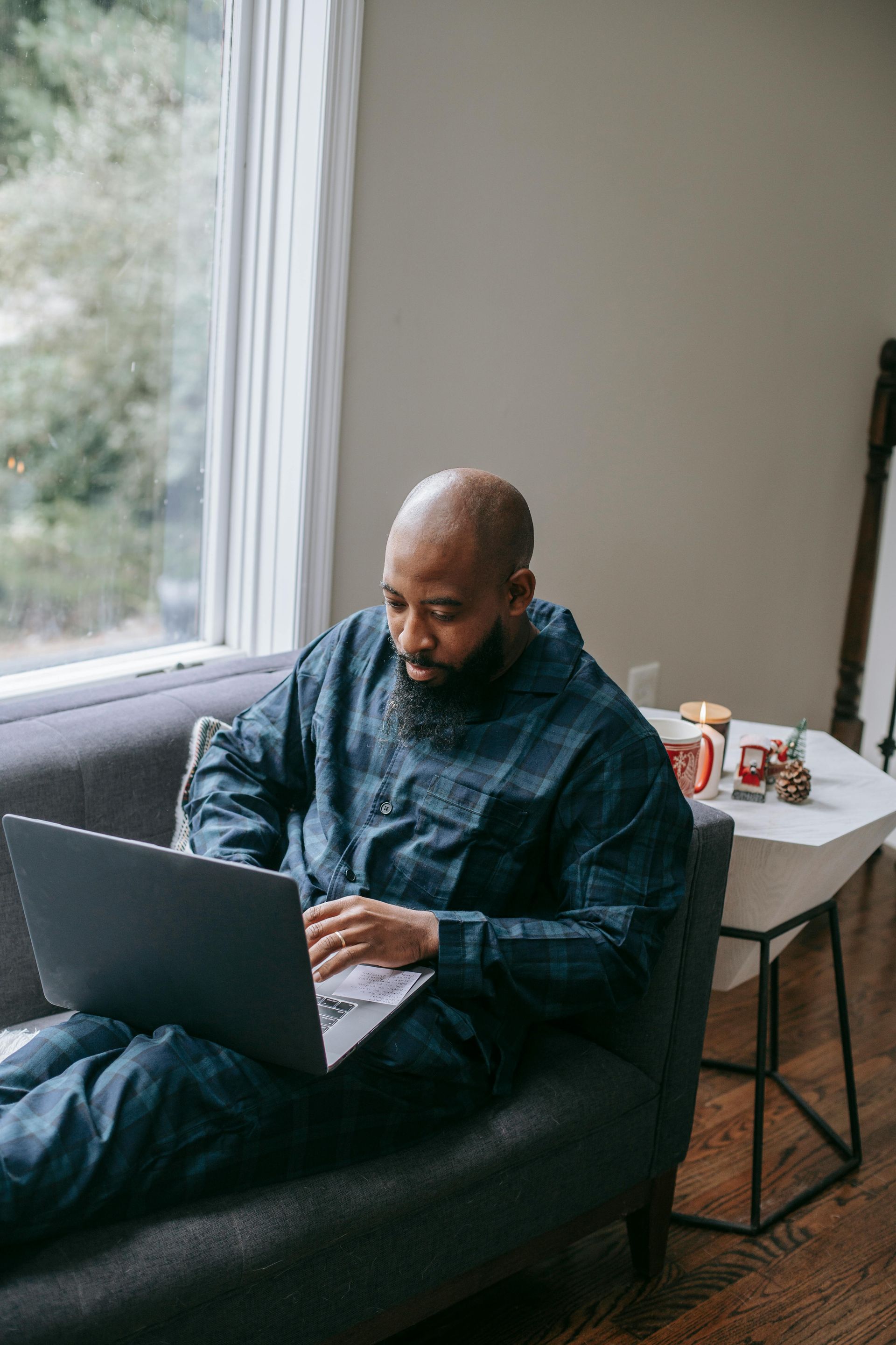 Man with a beard working on a laptop on a couch near a window.