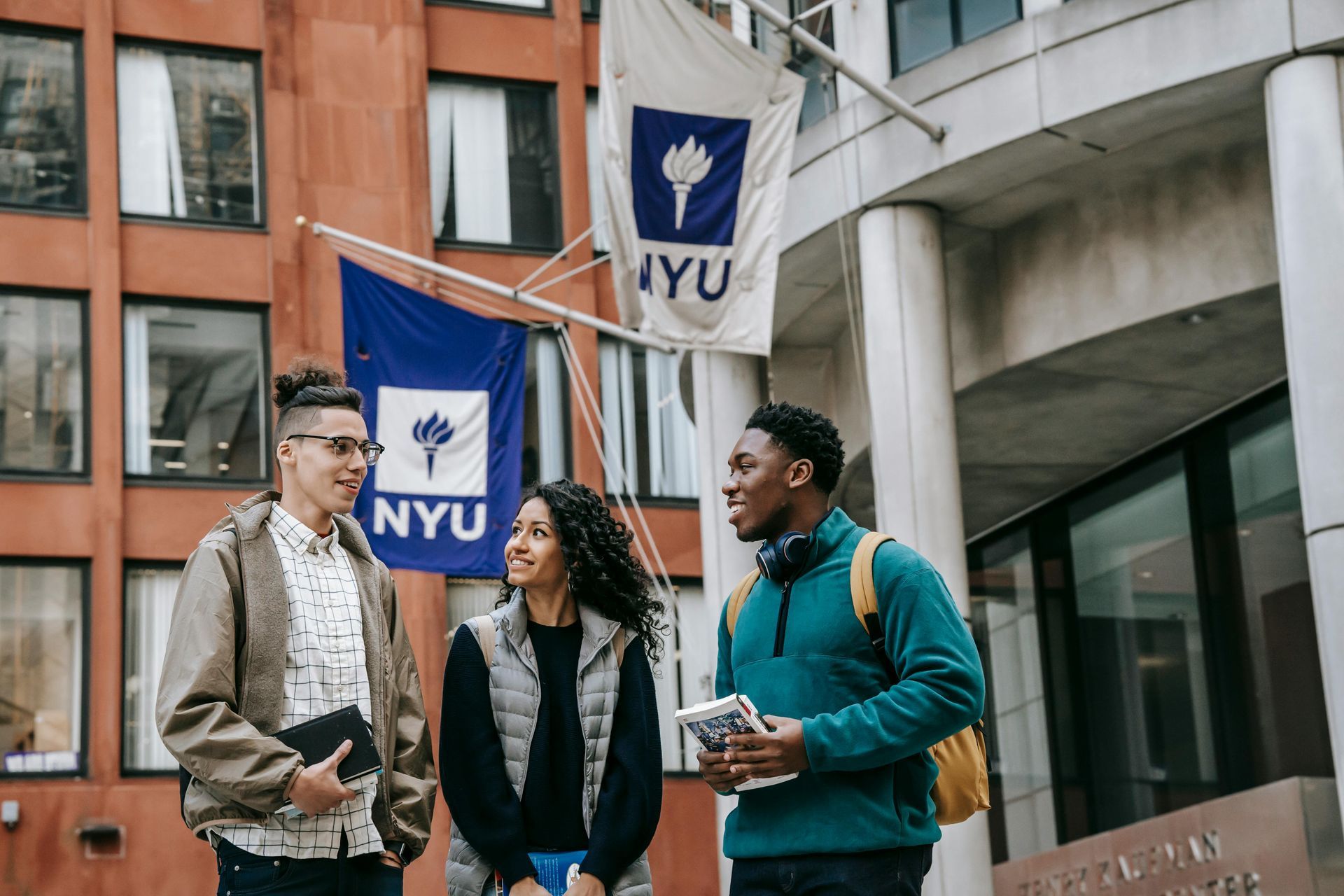 Three students conversing outside NYU building with NYU flags.