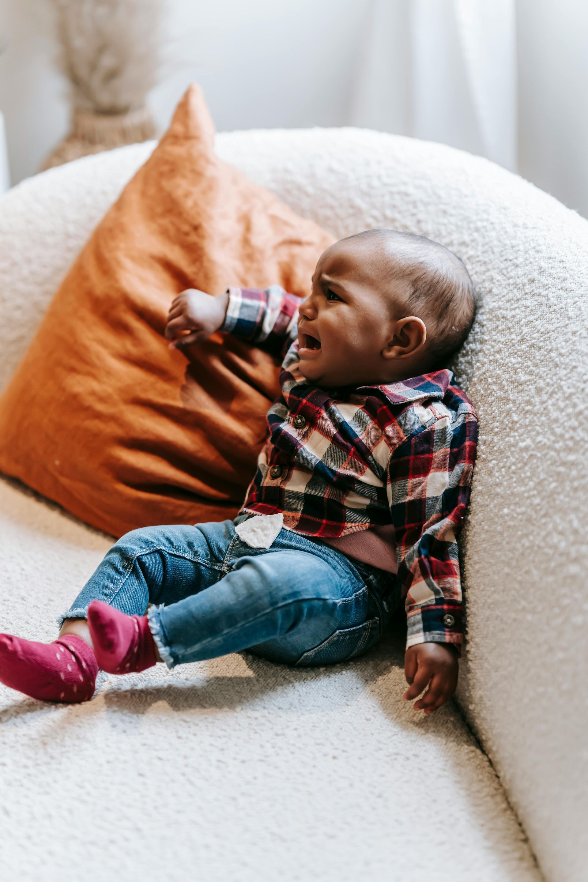 Baby in plaid shirt and jeans, crying on a white couch with an orange pillow. 