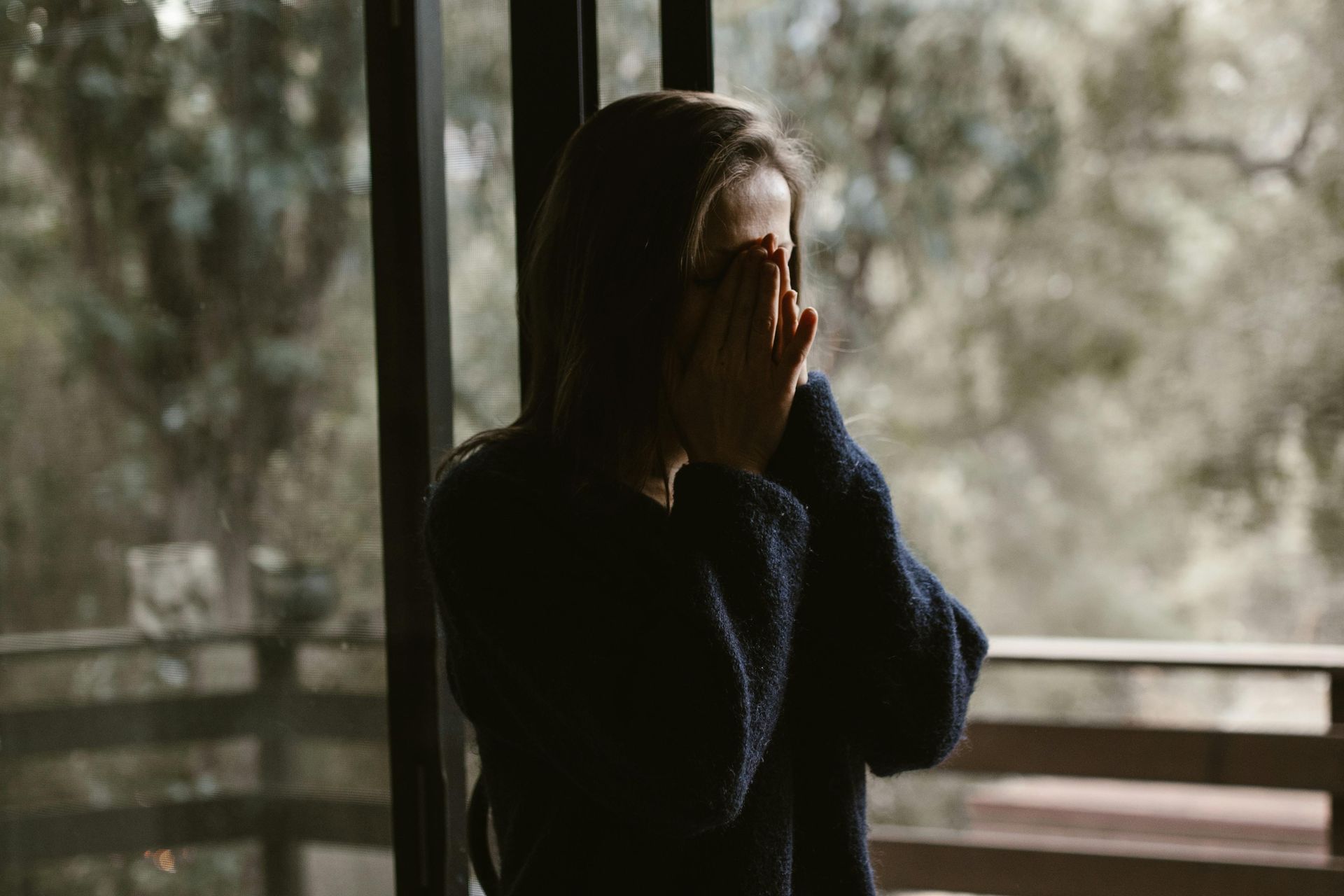 A woman is covering her face while standing in front of a window.