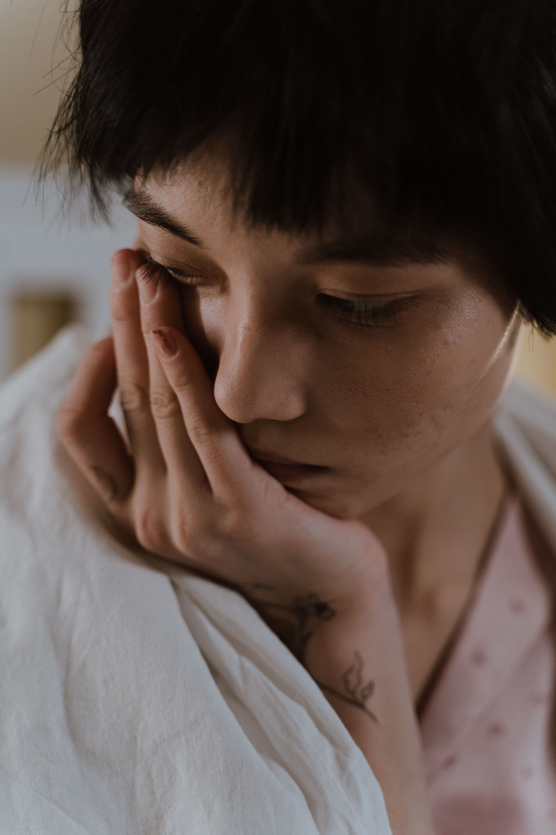 Person with short dark hair rests their face in their hand, looking down in a soft-lit close-up.