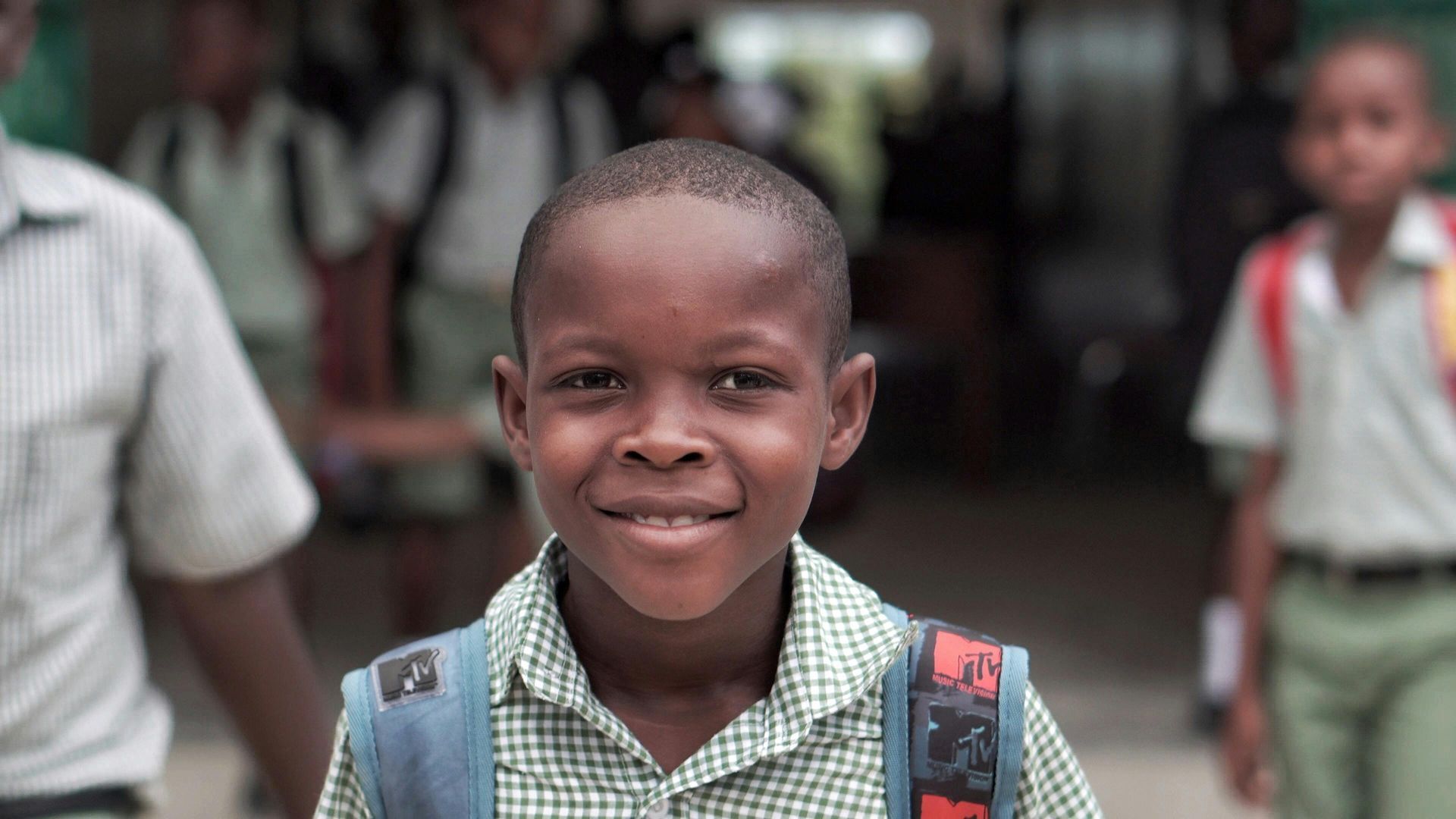 A smiling student in a green checkered shirt and backpack stands in a school hallway with other students in the background.