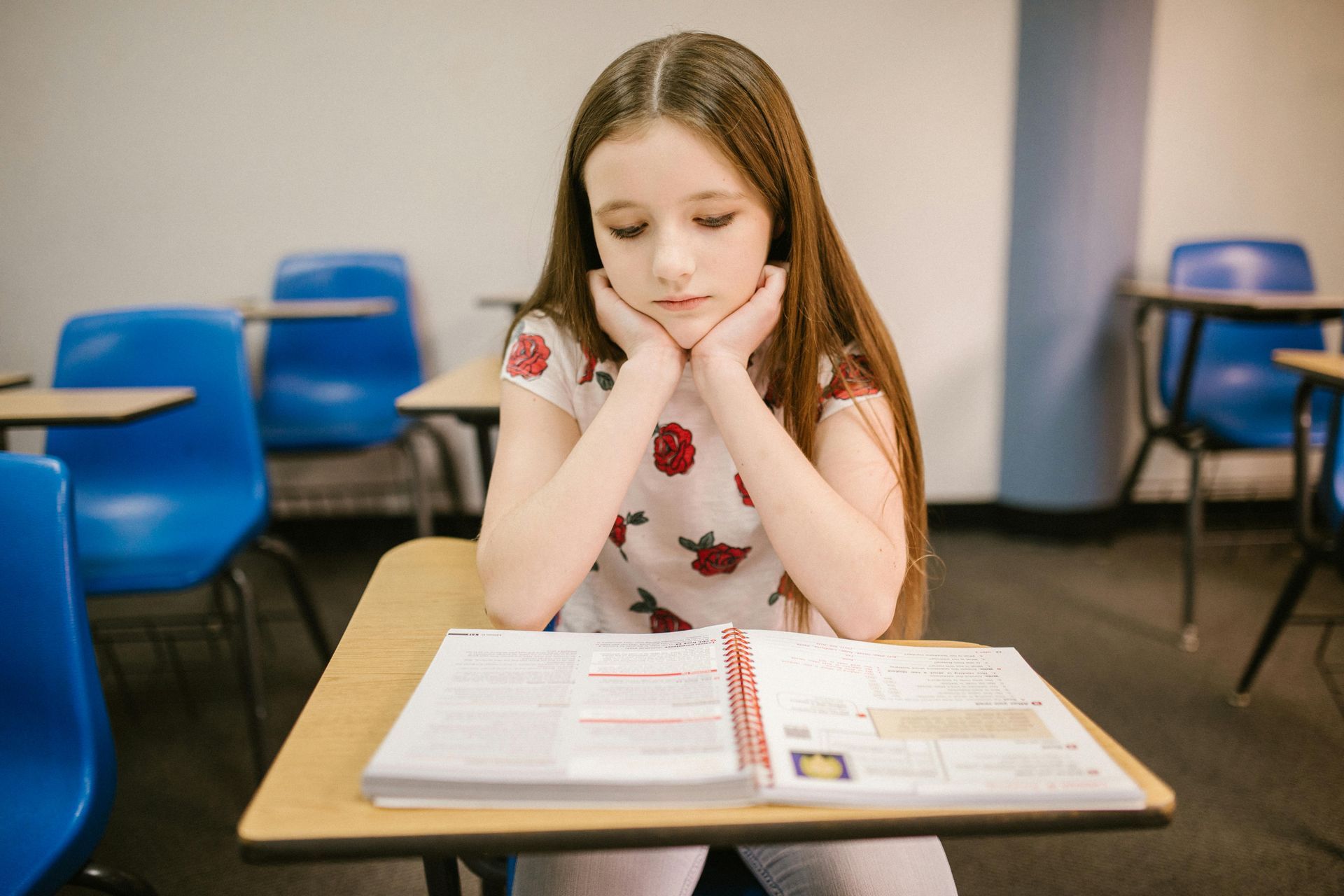 A young girl is sitting at a desk in a classroom with her head resting on her hands.