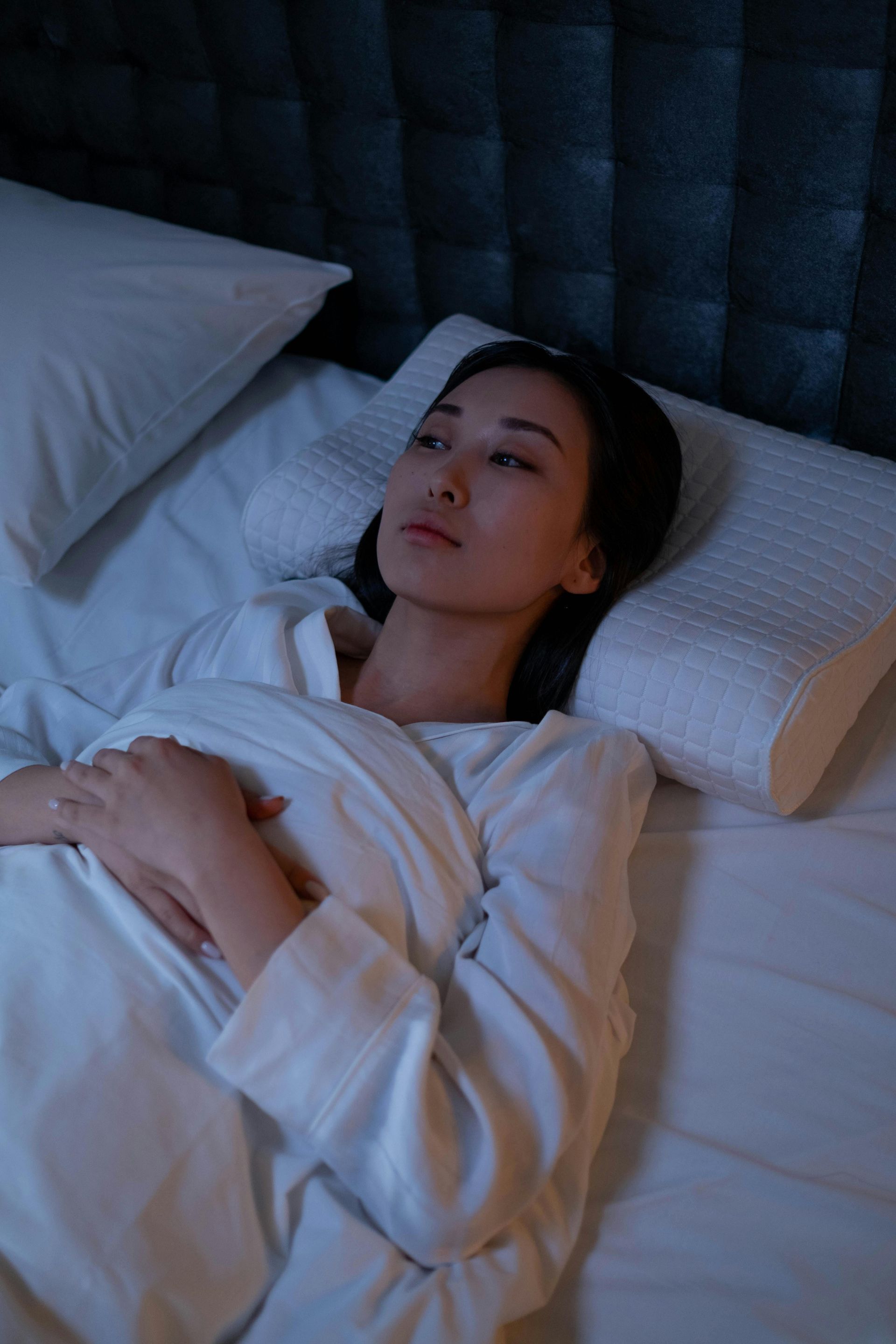 Person lying in bed under white blankets, staring upward in soft blue light.