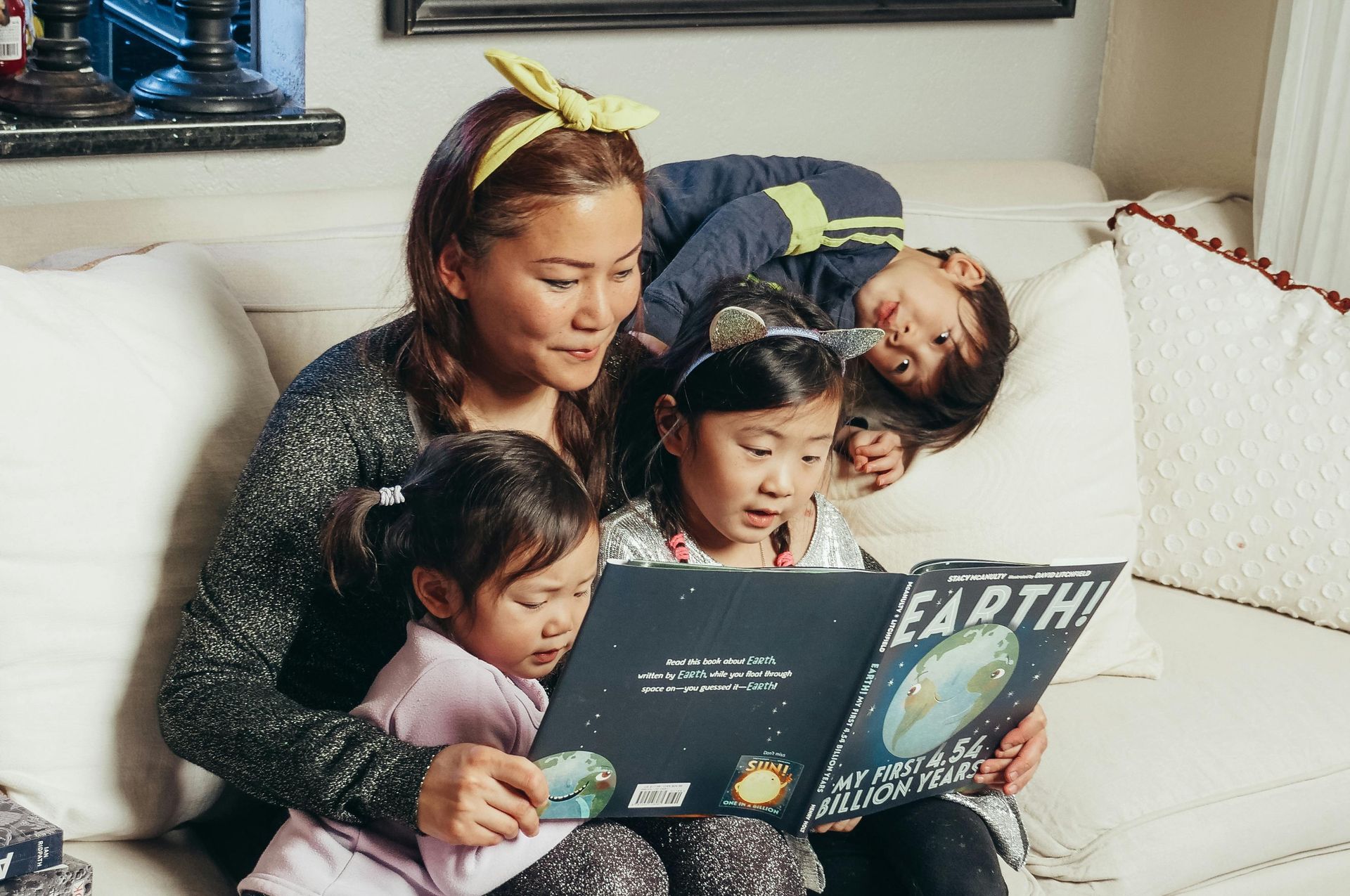 Woman reading a book to three children on a white couch. The book is about Earth.