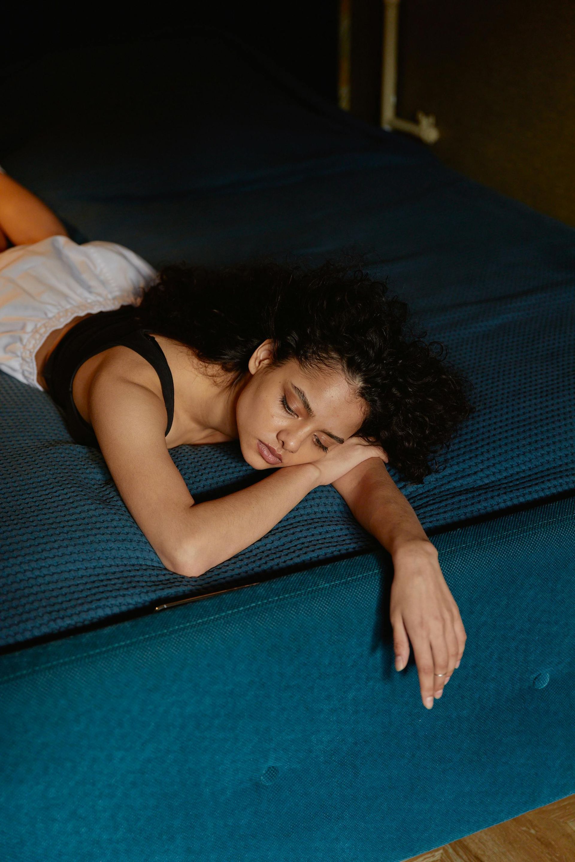 Woman with dark curly hair resting on a teal bed, wearing a black top and white shorts.