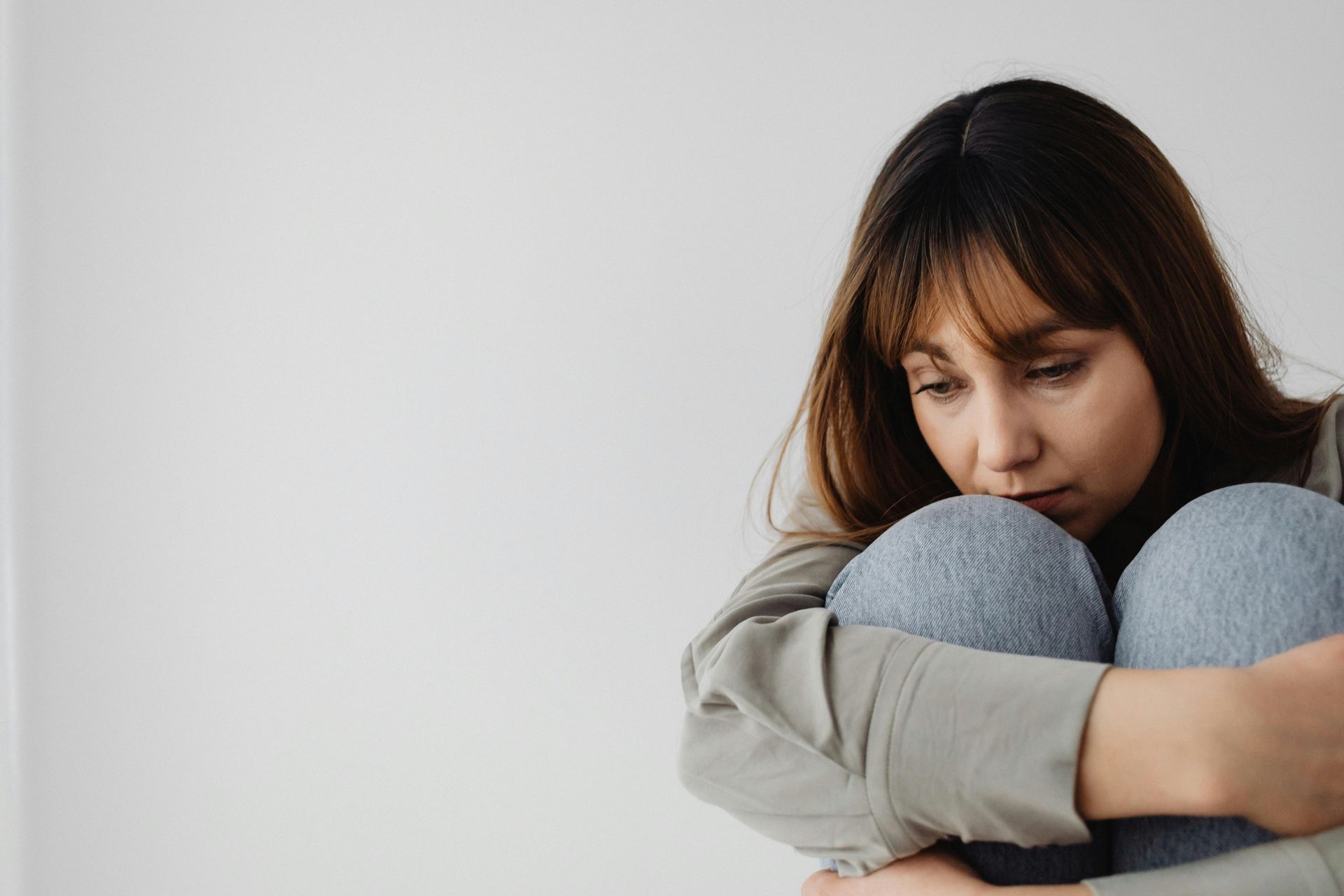 Woman sitting, hugging knees, looking down. Gray sweats, neutral background.