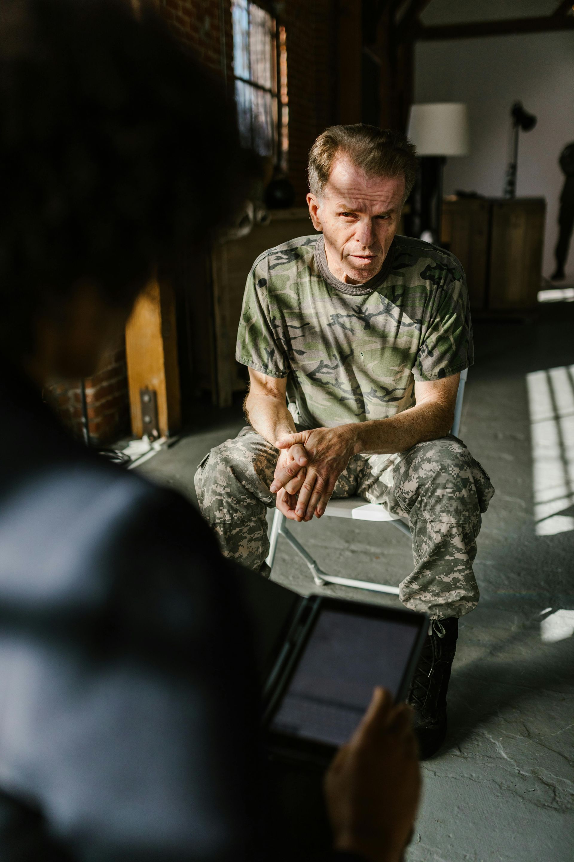A person in military fatigues sits talking to another person holding a tablet.