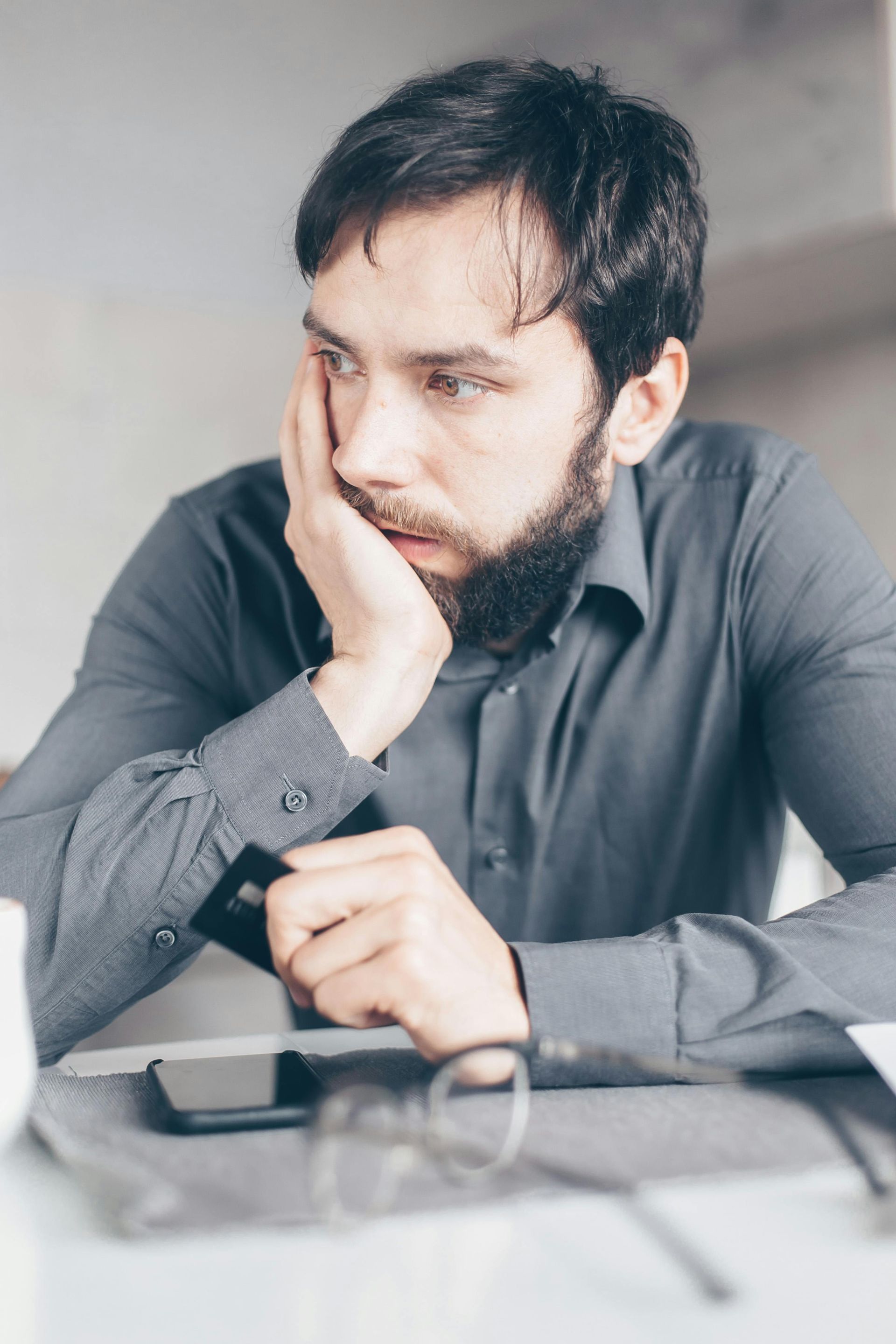 Man with beard resting his head on his hand, looking pensive. A phone, glasses, and credit card are visible on the table.