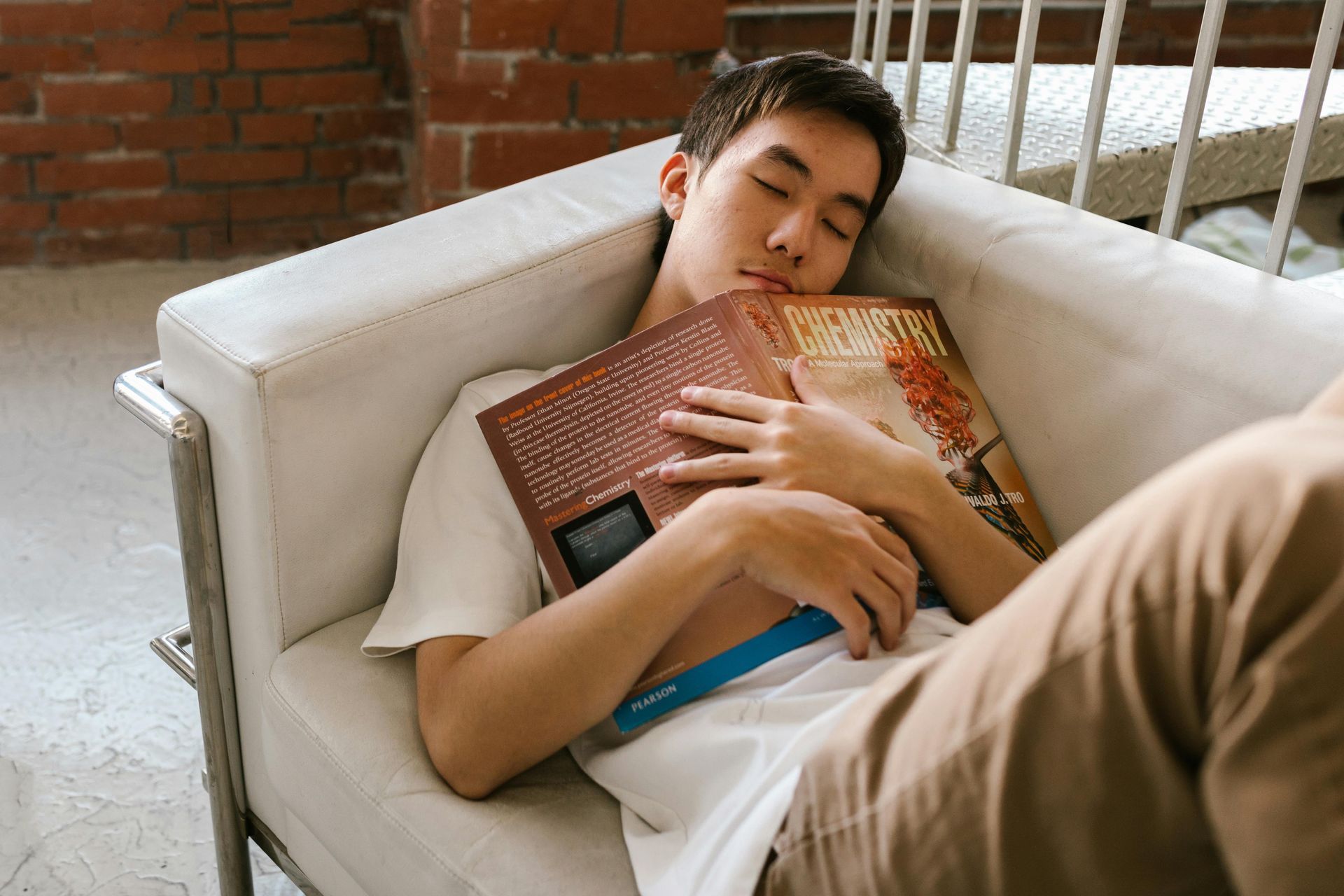 A teenager sleeps on a white couch, hugging a book. Wearing brown pants and a white t-shirt.