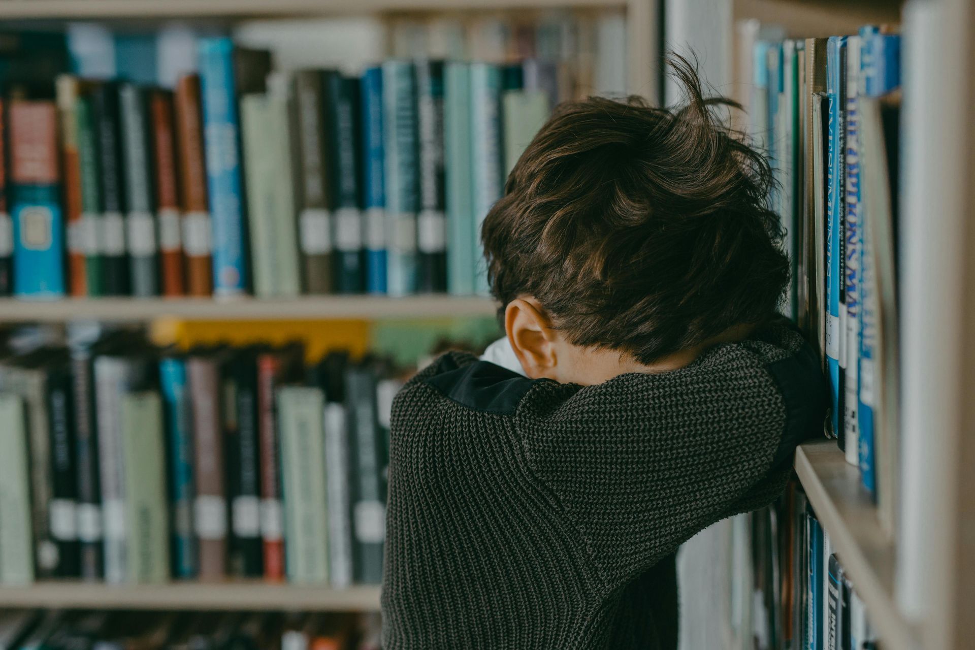 A young boy is leaning against a bookshelf in a library with his head in his hands.