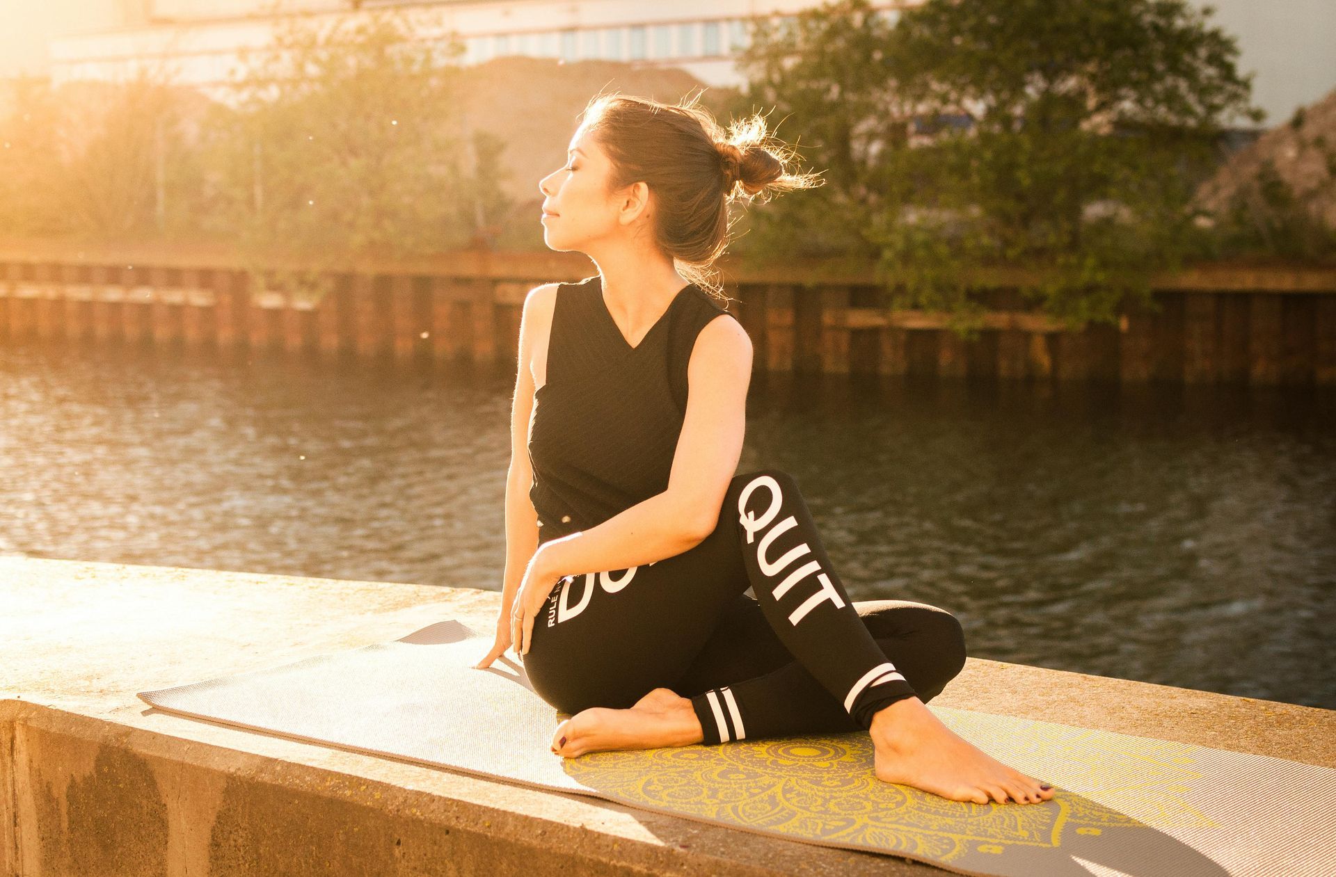 Woman in black athletic wear twists, sitting on a yoga mat by water, with a sunny backdrop.