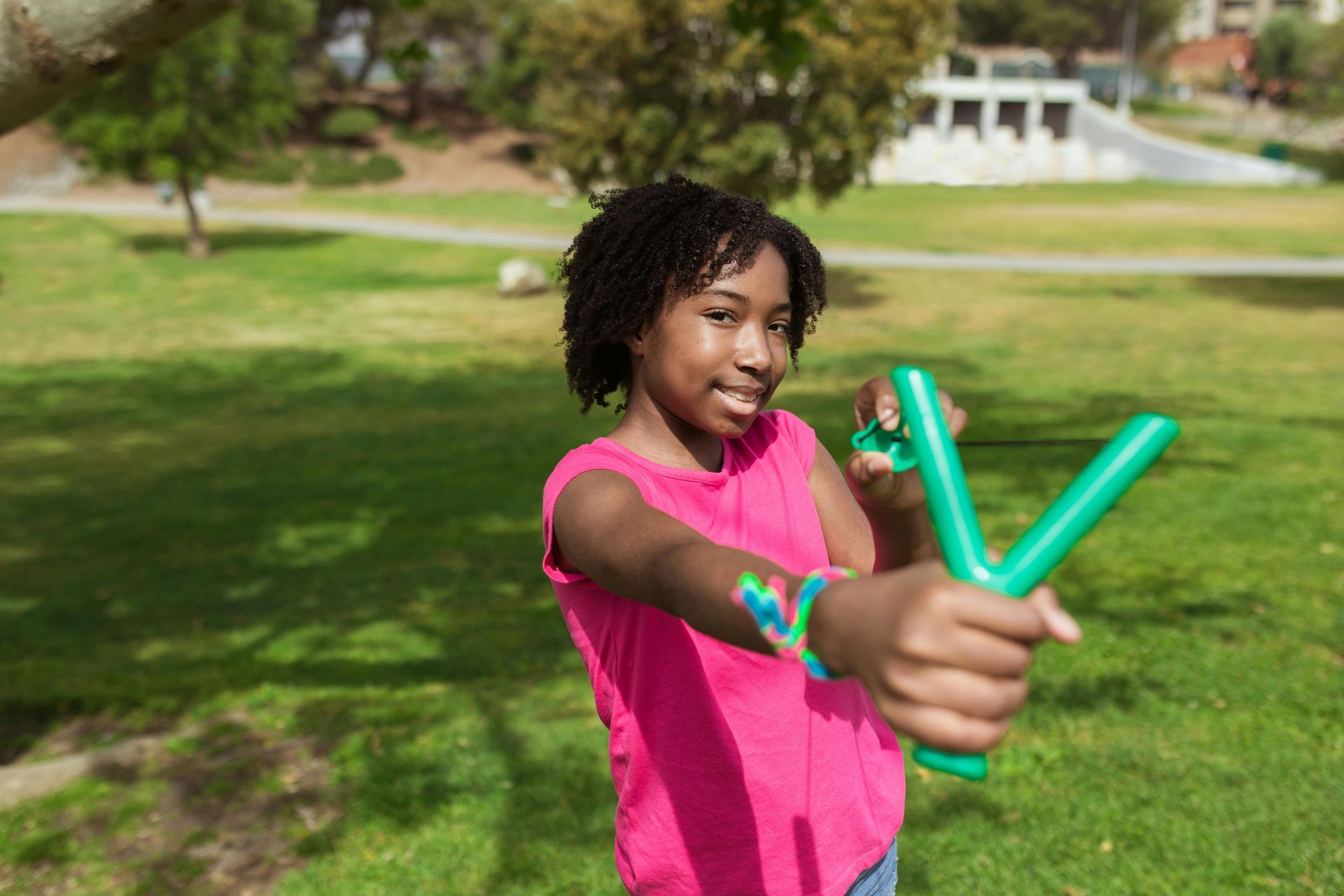 Girl aims a green slingshot in a park, wearing a pink shirt, on a sunny day.