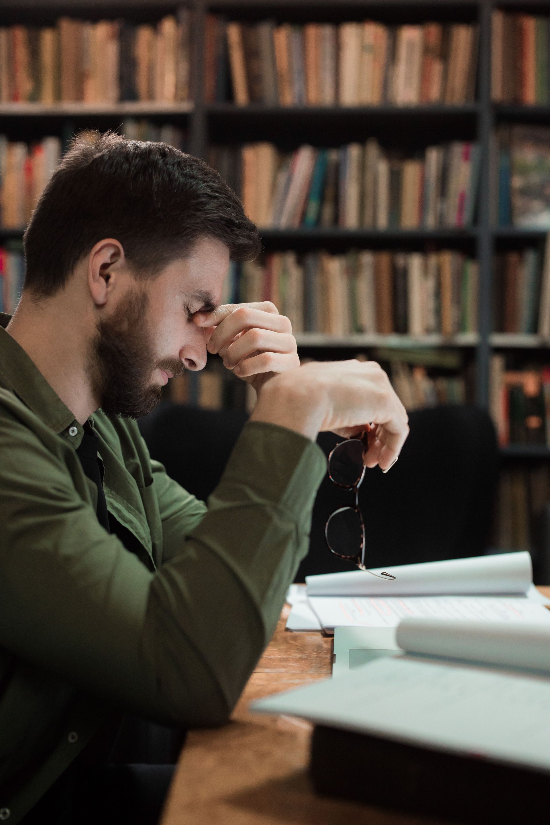 Man sitting at a desk in a library, reading with a hand on his face and bookshelves in the background