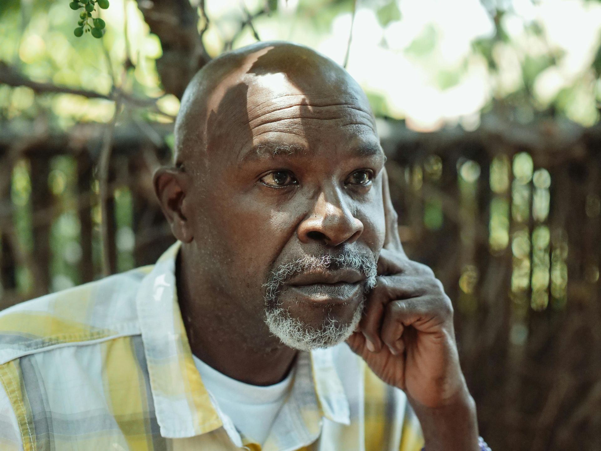 A thoughtful senior man with a partly gray goatee, resting a hand on their face, in a sunny outdoor setting.