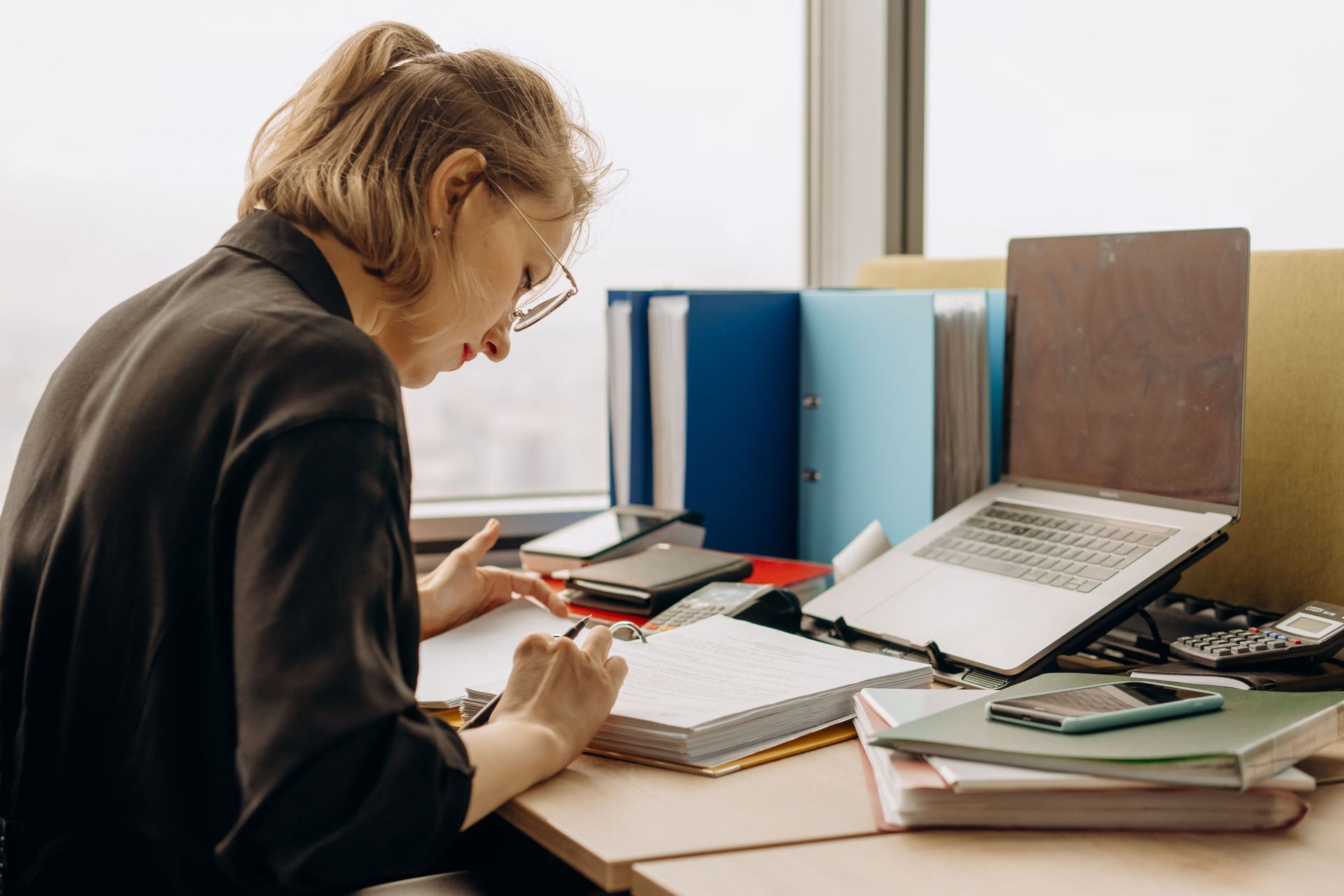 Person writing at a cluttered desk with books, folders, and an open laptop in a bright office.