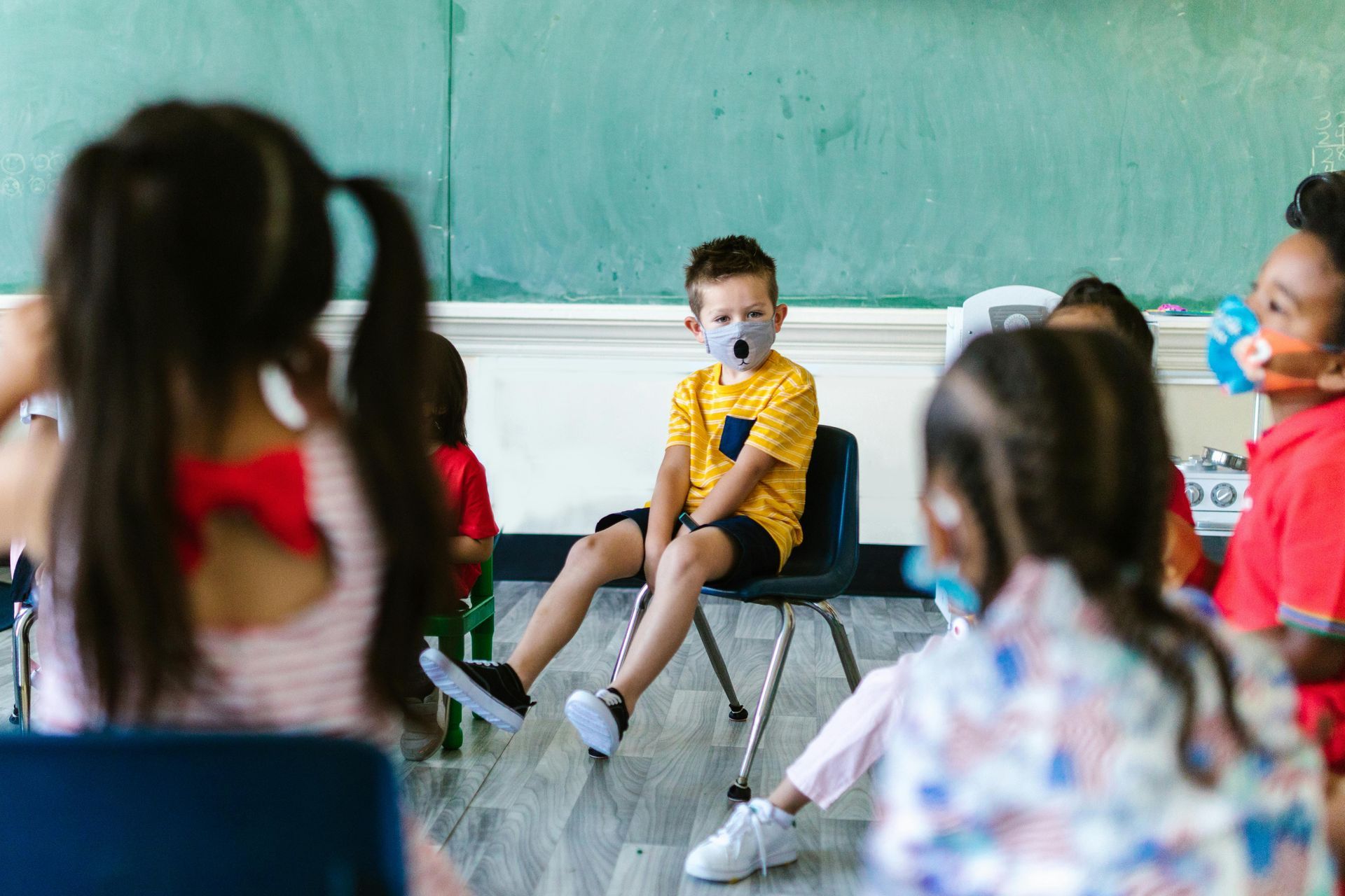 A group of children sit in a classroom, with one child in a yellow shirt and face mask seated in the center.