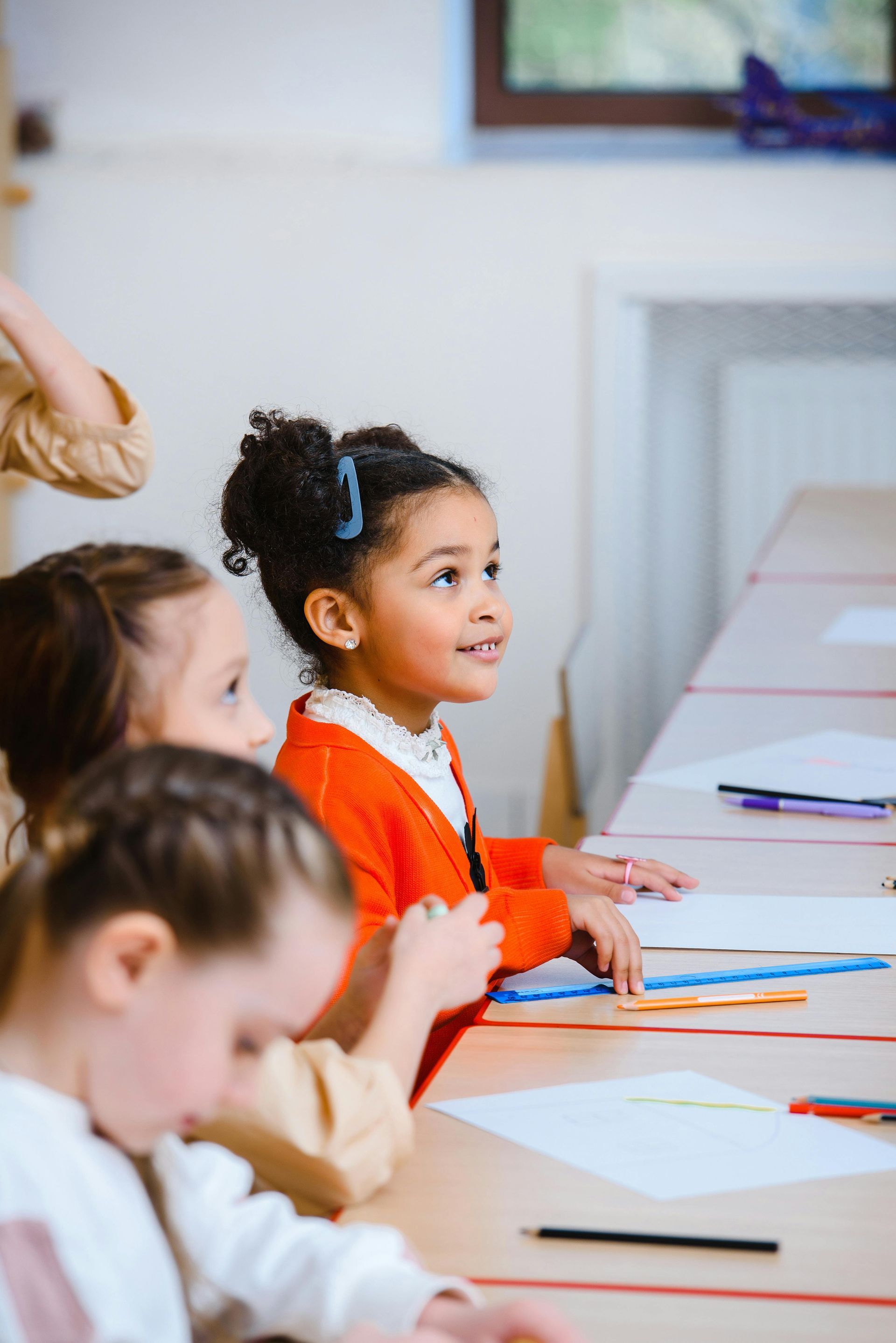 Students sit at desks in a classroom, looking forward, with papers, rulers, and pencils on the table.