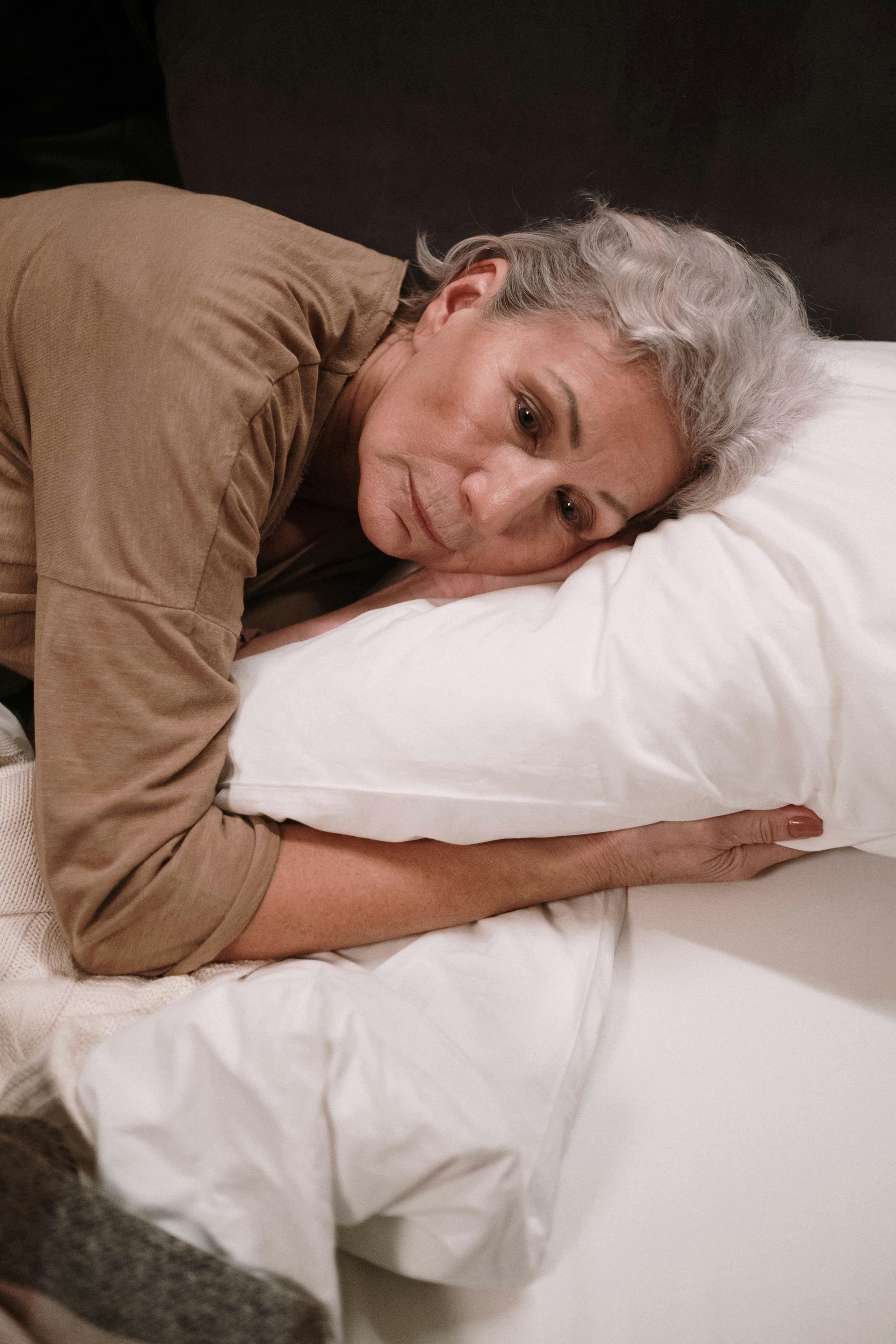 Woman resting head on white pillow, looking down with a somber expression. Brown shirt, gray hair, indoors.