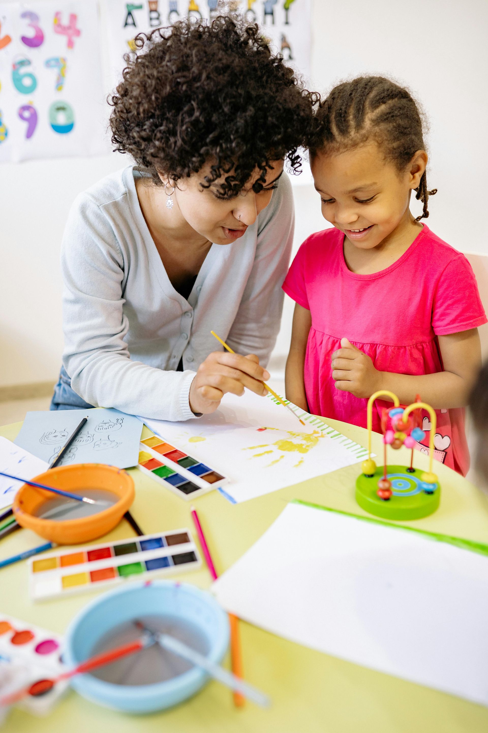 A woman helps a girl paint with watercolors at a table in a classroom.