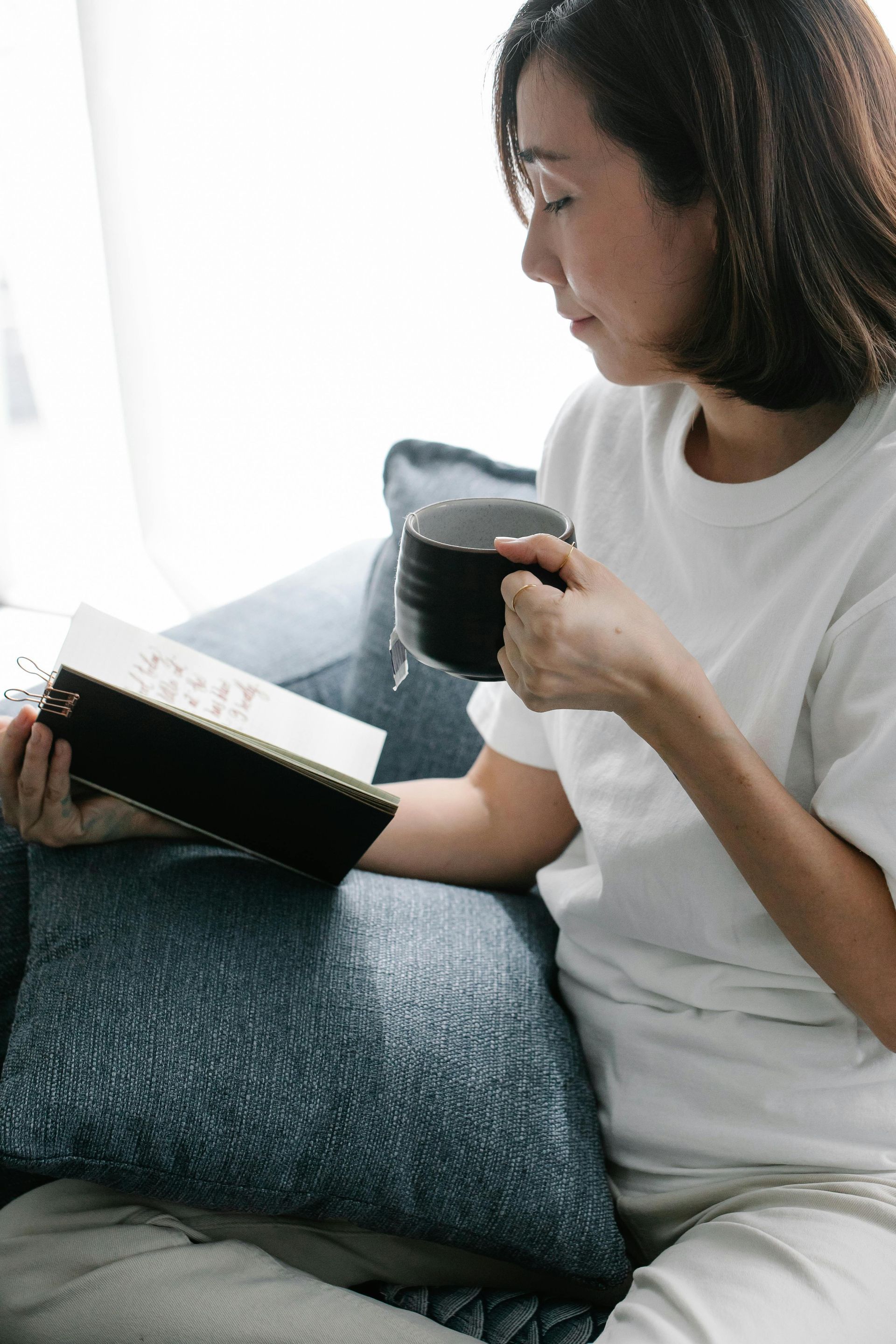 Woman reading a book, holding a coffee mug, seated on a couch near a window.