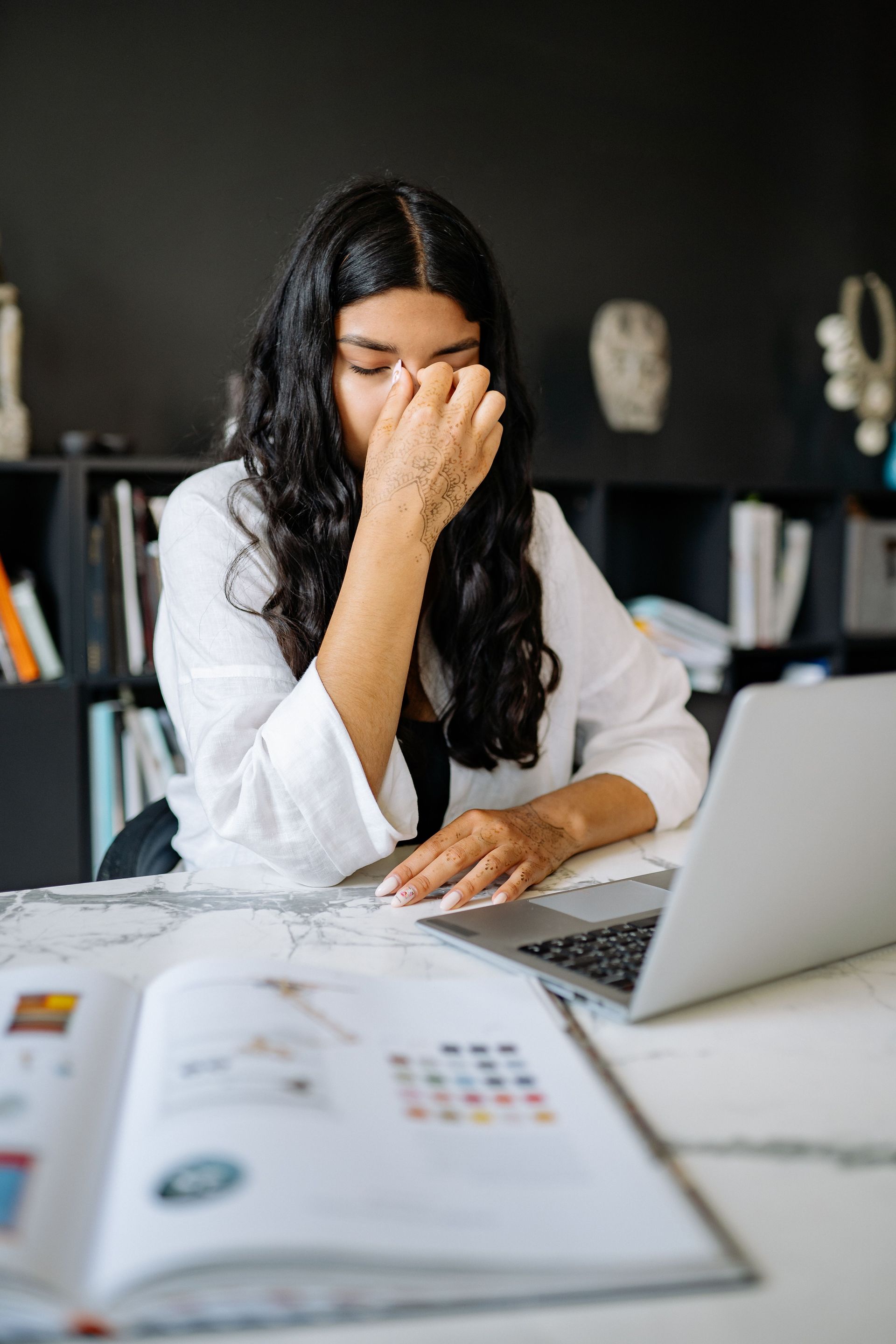 Person at a desk rubbing their eyes while working on a laptop, with papers and a notebook in front.