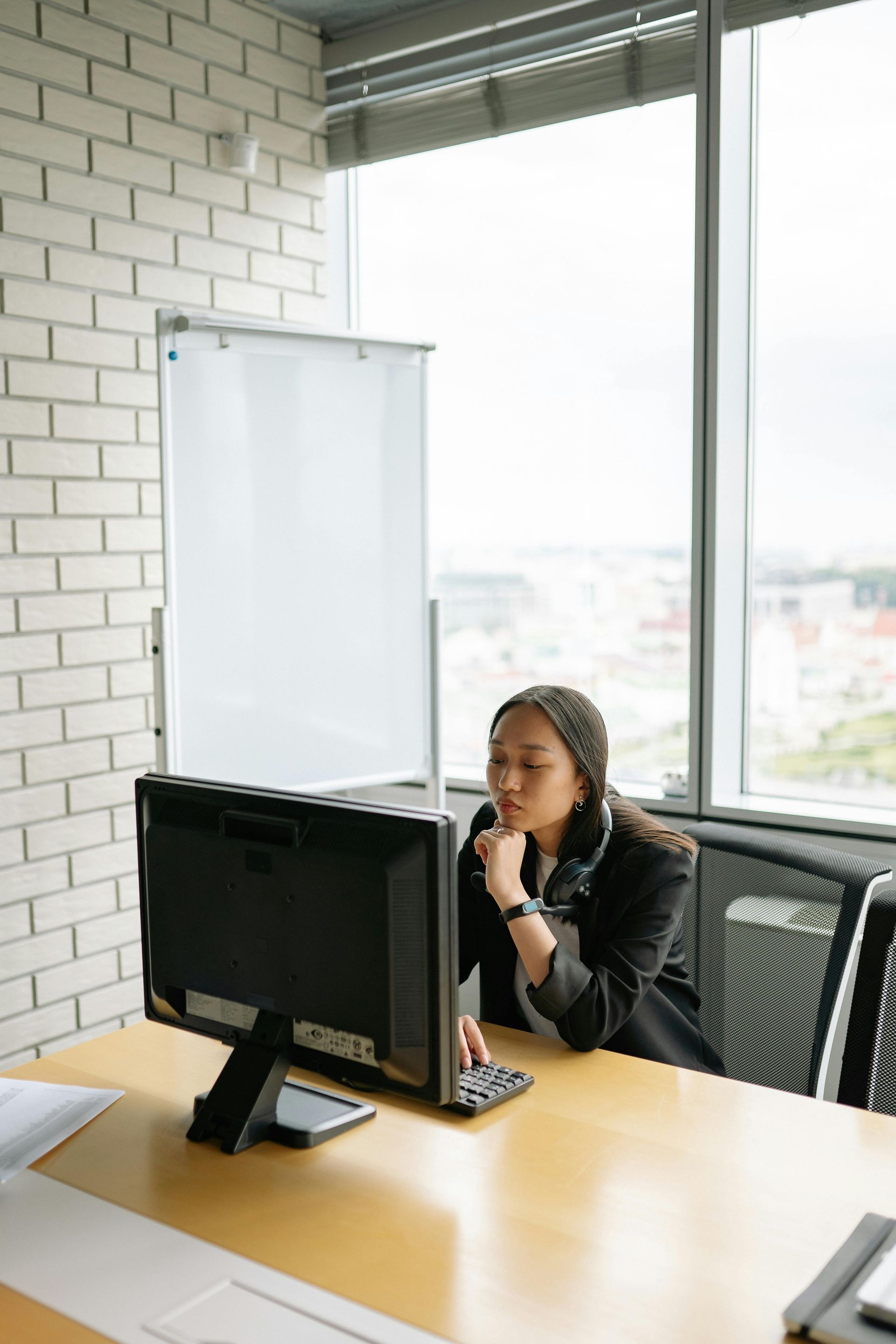 Woman at computer in office, thinking, hand on chin. Whiteboard and city view in background.