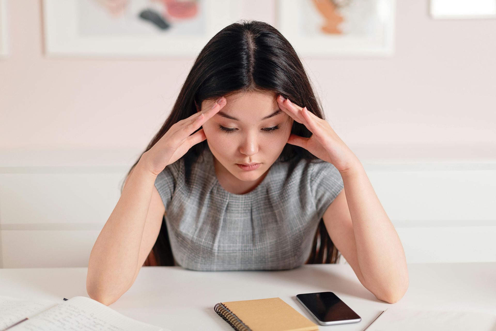Woman with long dark hair, hands on head, looking down at desk with notebook and phone.