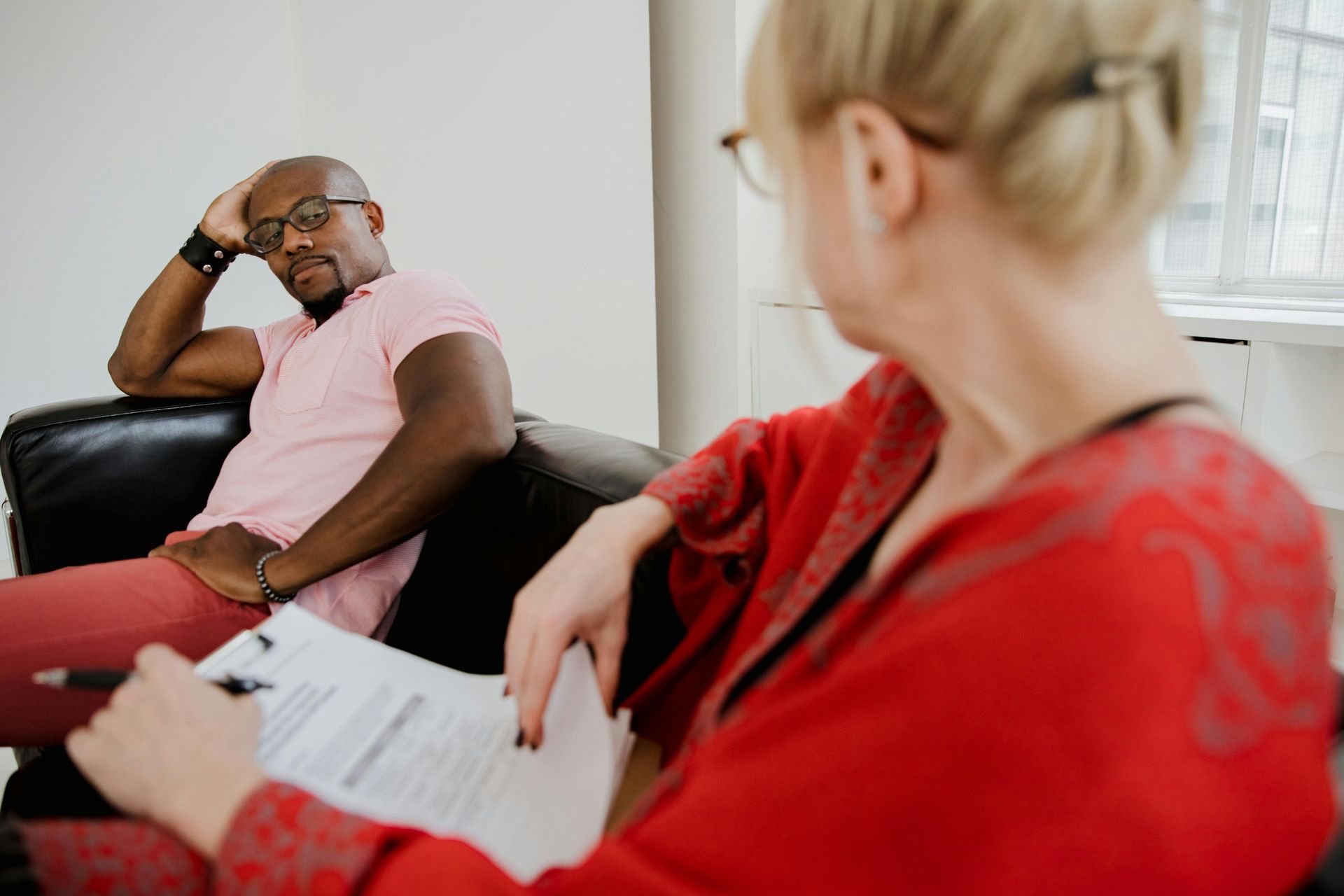 Man in therapy, leaning on arm, facing a therapist in a red top, holding a clipboard.