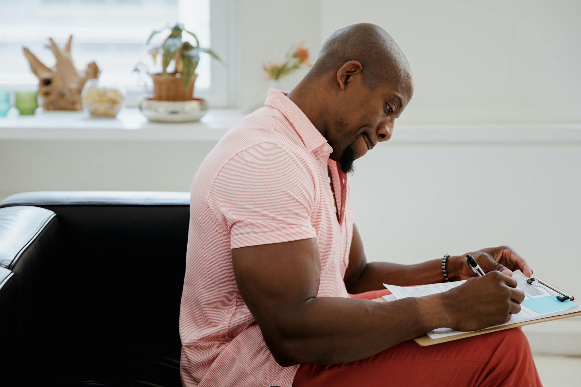 Man in pink shirt writing on a clipboard, seated on a black couch indoors.