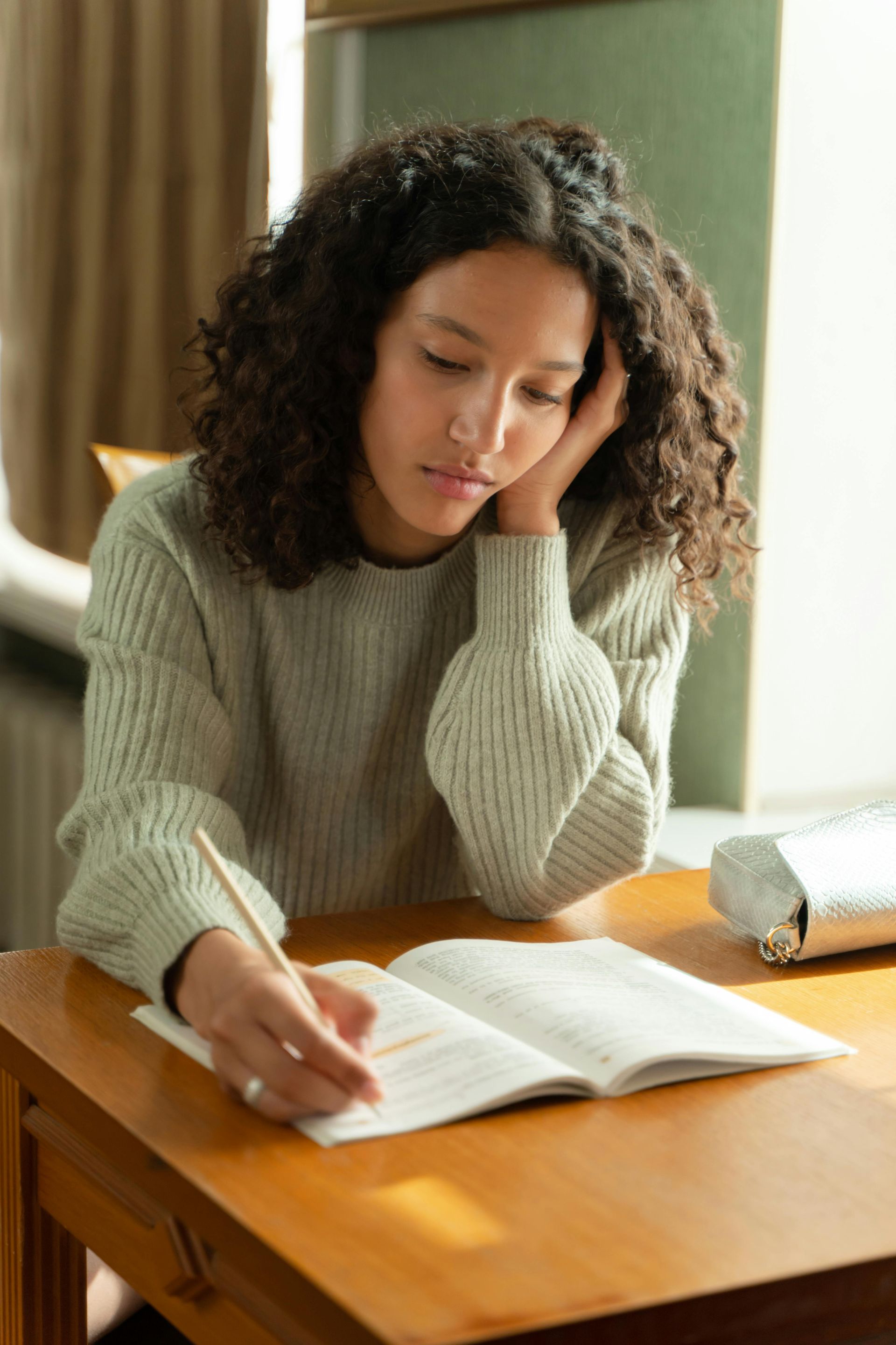 Teen with curly hair writing in a notebook at a wooden table, looking down with a hand on her cheek.