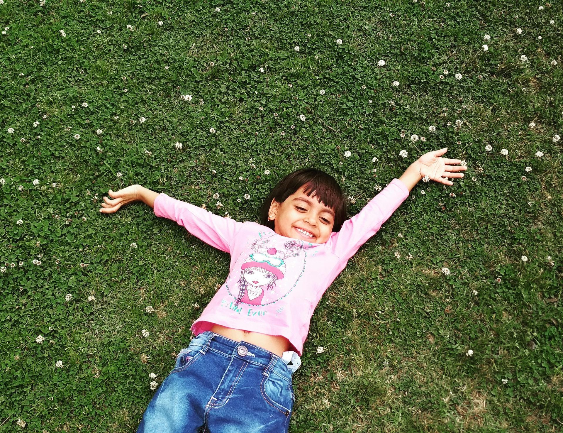 A little girl is laying on the grass with her arms outstretched.