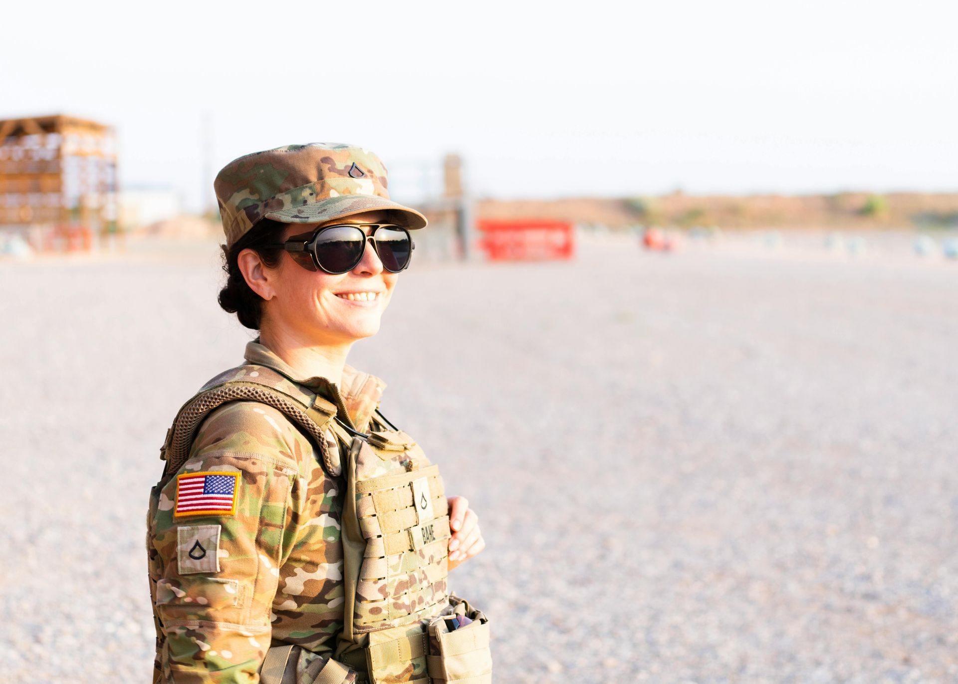 Soldier in camo uniform and sunglasses smiles outdoors, wearing body armor, desert setting.