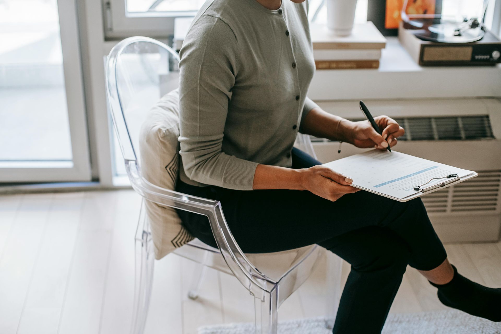 Woman in grey sweater and black pants writing on a clipboard while sitting in a clear chair.