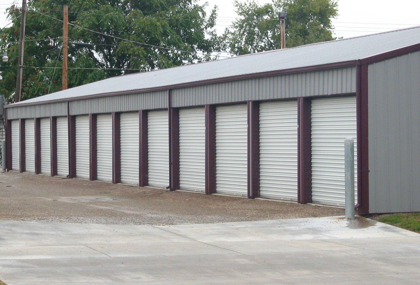 Row of gray storage units with red-brown frames and white roll-up doors under a metal roof.
