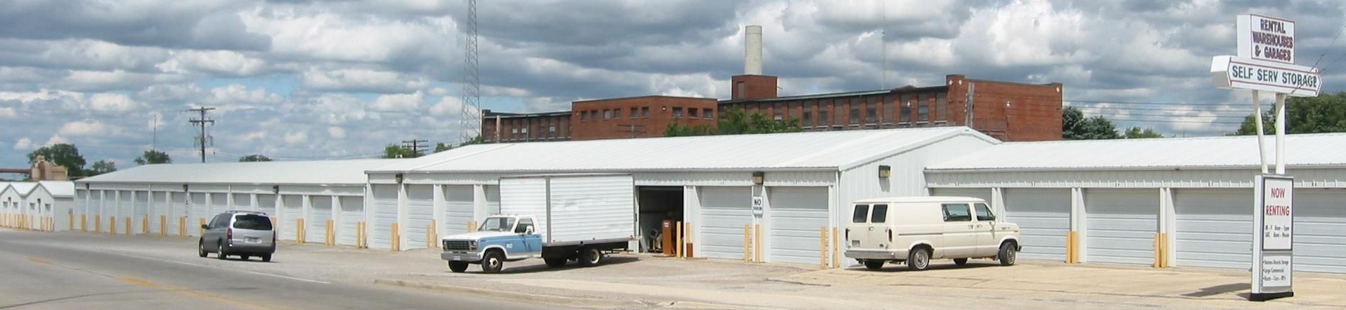 Storage units with vehicles in front, including a truck, a van, and a car. A brick building is in the background.