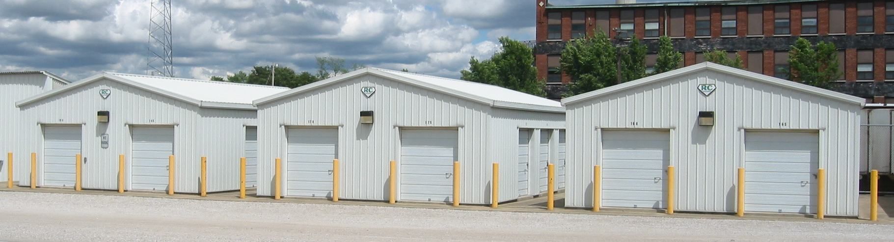 White storage units with roll-up doors in a row, with a cloudy sky and a brick building in the background.