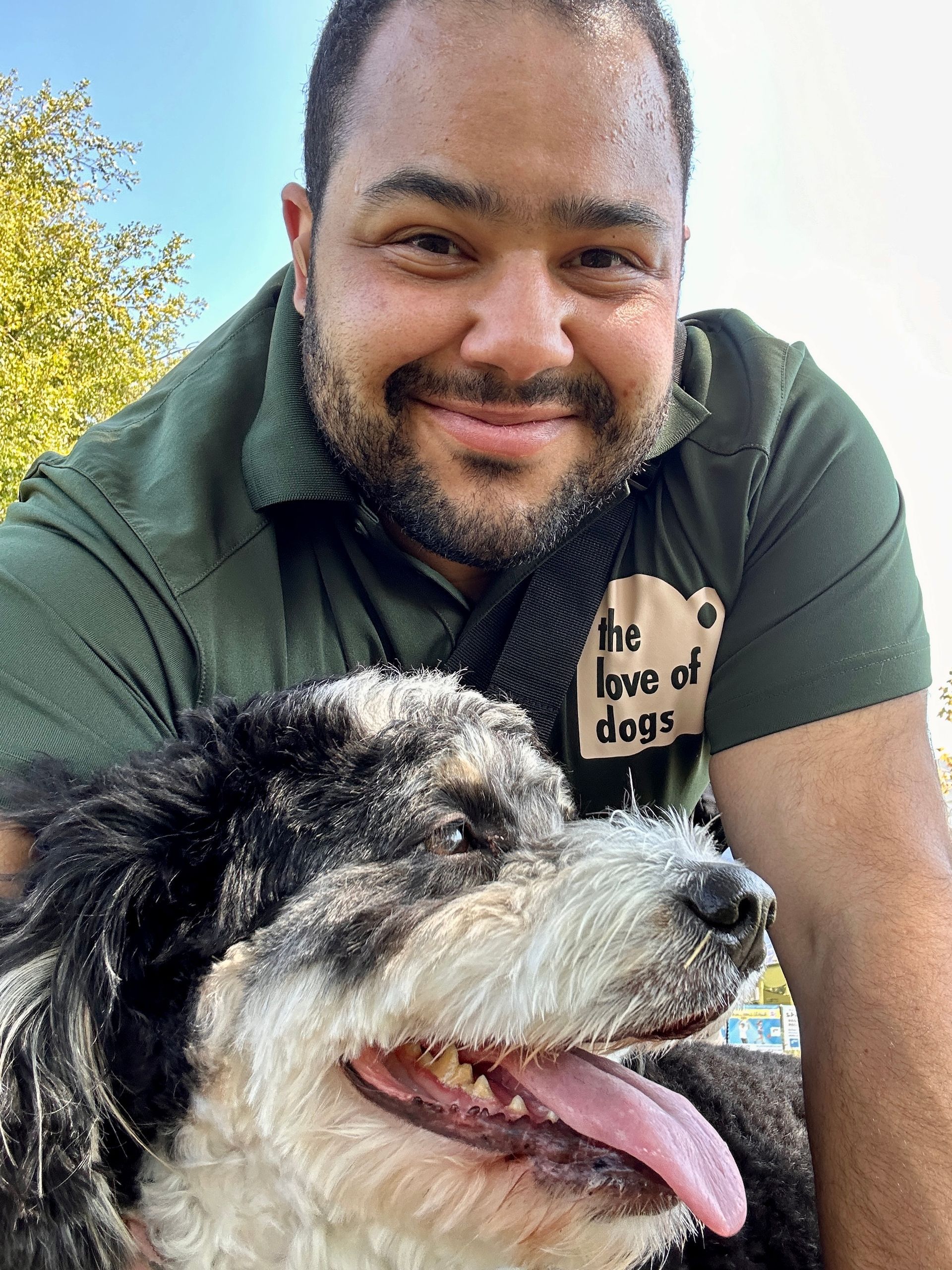 Man smiling with a black and white dog outdoors; wearing a green shirt that says