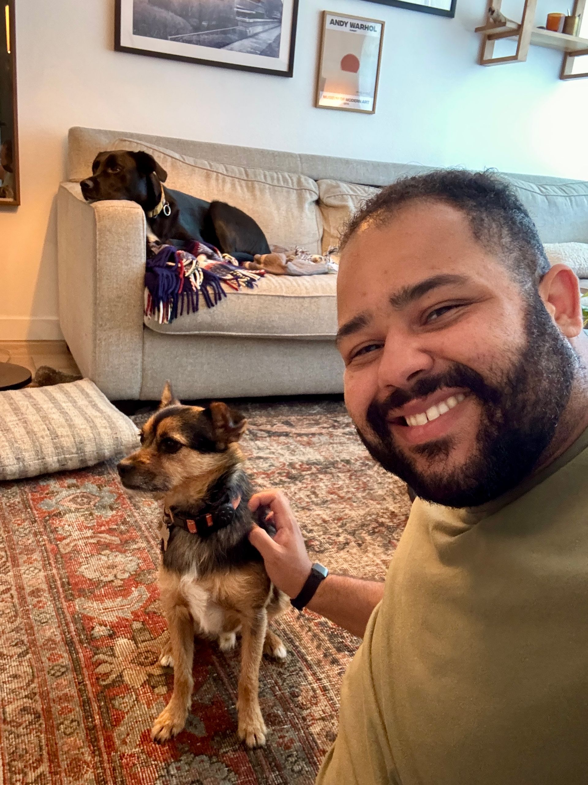 Man smiles, petting small dog on patterned rug; black dog rests on couch in background.