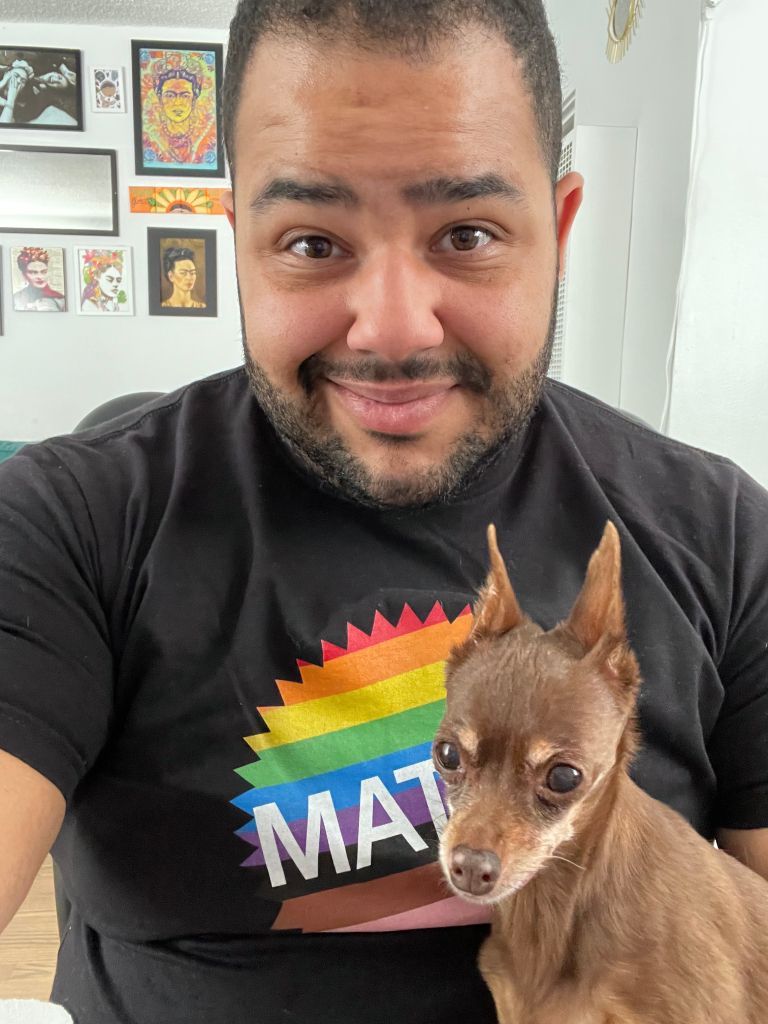 Man smiling, holding small brown dog. Man's shirt has rainbow design and 